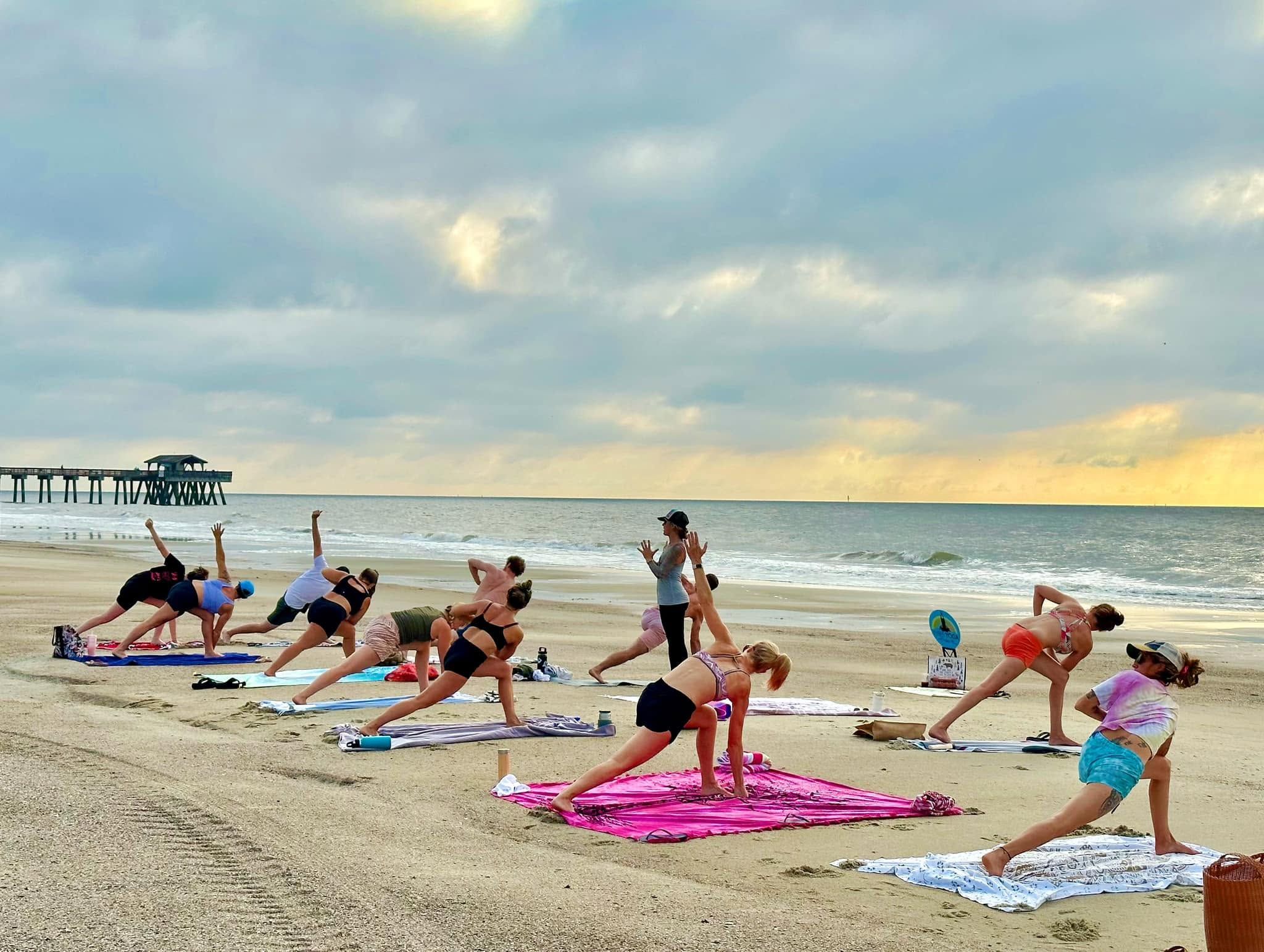 A group of people practice yoga on colorful towels and mats spread across the sand, holding a side-angle stretch as an instructor leads the class near the Tybee Island Pier. Soft morning light breaks through clouds over the calm ocean behind them. 