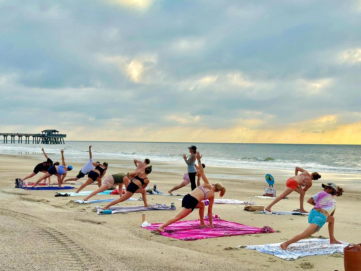 A group of people practice yoga on colorful towels and mats spread across the sand, holding a side-angle stretch as an instructor leads the class near the Tybee Island Pier. Soft morning light breaks through clouds over the calm ocean behind them.