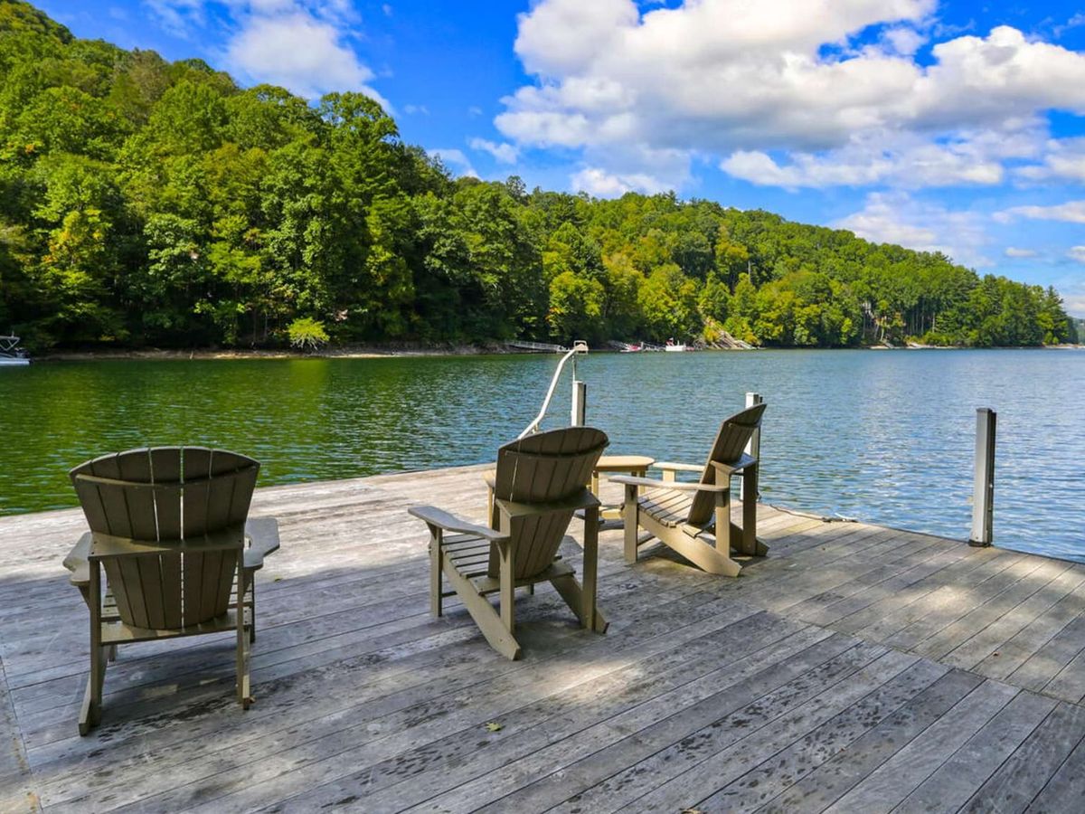 Two Adirondack chairs sit on a private dock overlooking a calm mountain lake surrounded by green hills. The peaceful water and blue sky make it the perfect place to relax, sip coffee, or watch the sunset. This quiet lakefront retreat is ideal for a North Carolina mountain getaway.