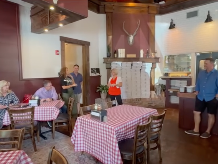 A cozy restaurant interior with red-and-white checkered tablecloths, wooden furniture, and stacked pizza boxes features several people casually gathered and chatting near the counter and seating area.