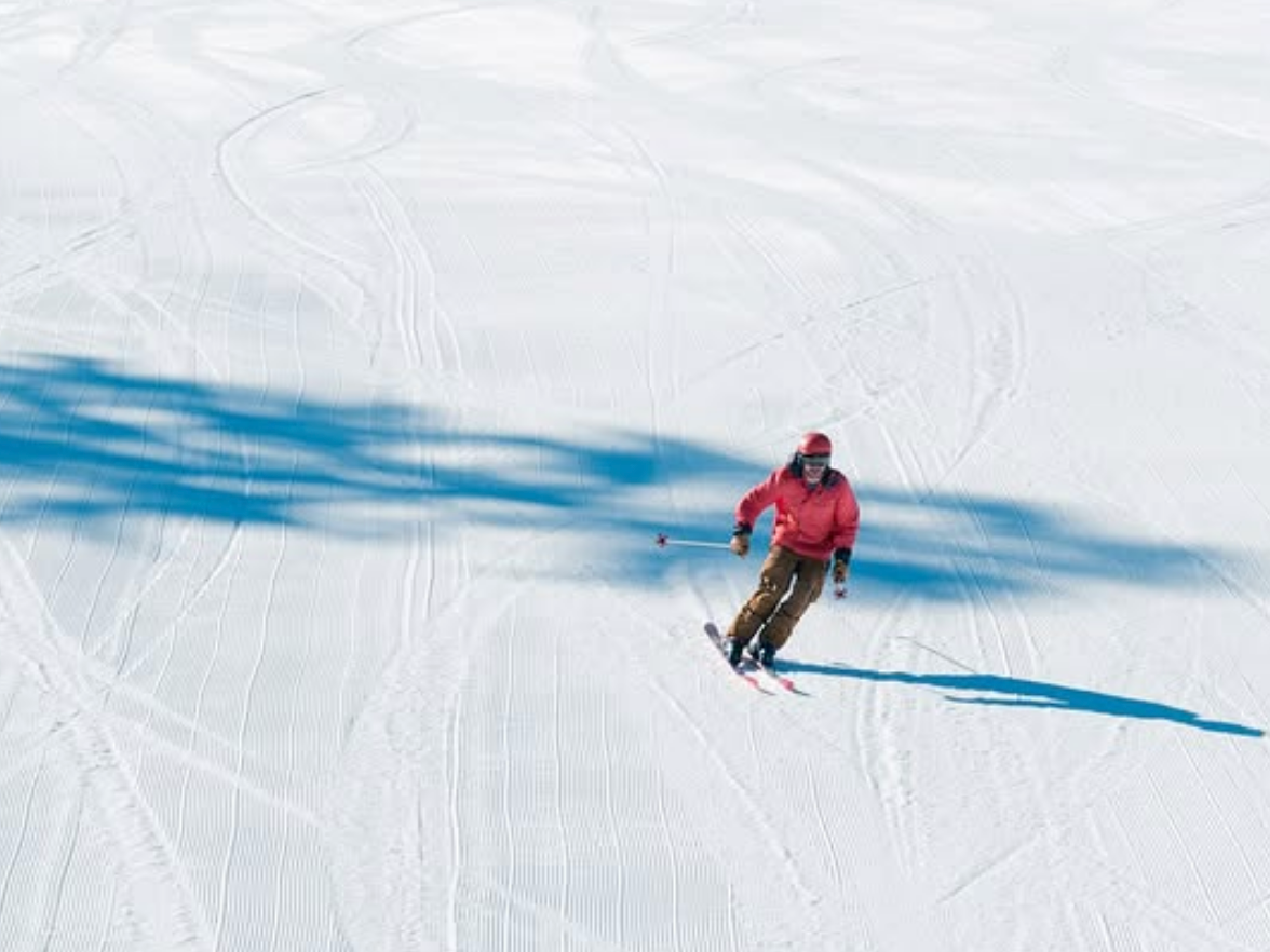 A skier in a red jacket carves down a freshly groomed snowy slope, casting a sharp shadow beneath a clear blue sky.