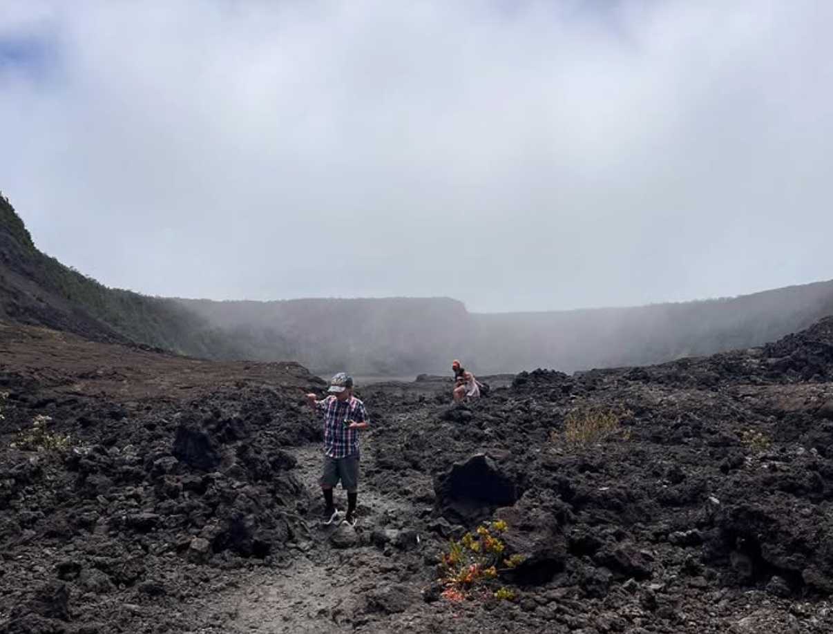 Hikers walk across the rocky floor of Kīlauea Iki Crater, surrounded by dark volcanic rock and mist rising from the rim in Hawai’i Volcanoes National Park.