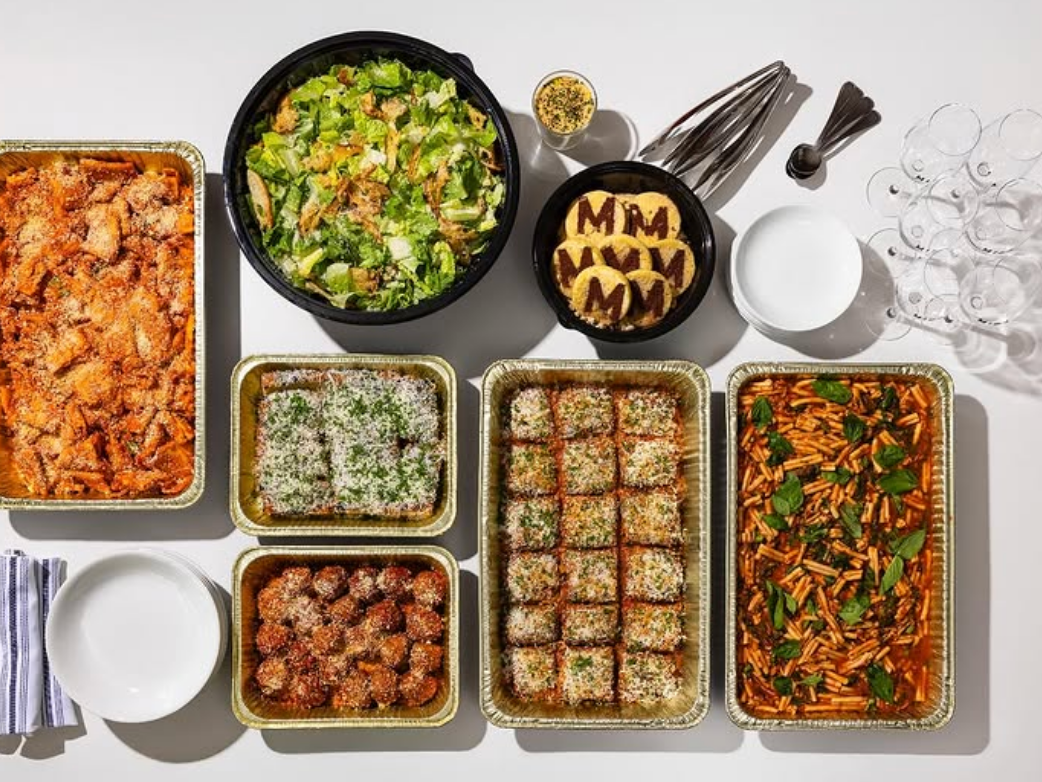 A vibrant catering spread featuring trays of pasta, lasagna, meatballs, salad, and desserts, all garnished with fresh herbs and cheese, arranged neatly on a white table alongside plates, utensils, and wine glasses.