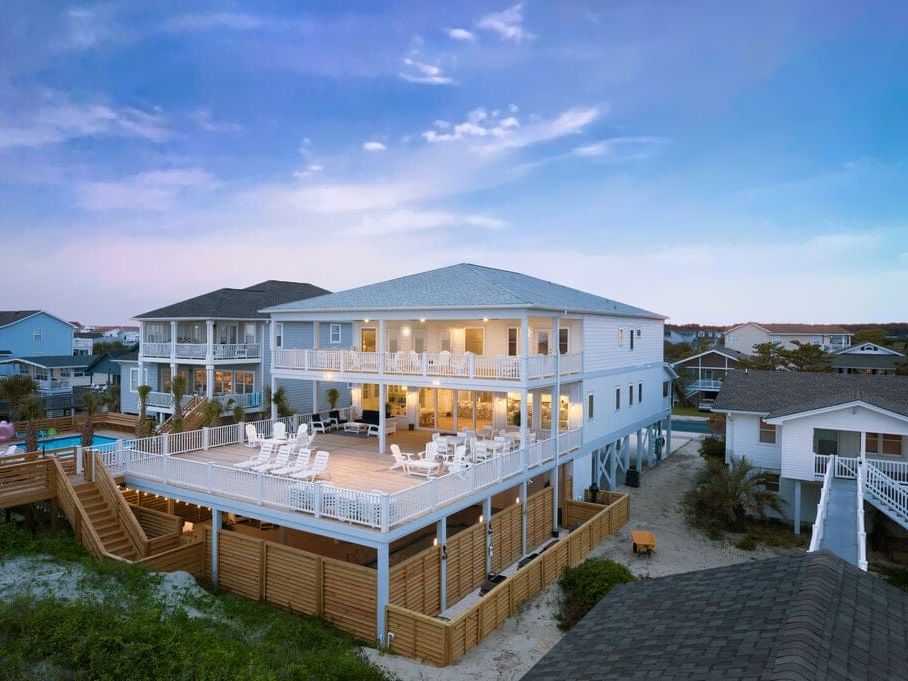 A large, multi-story beach house with expansive decks, white railings, and outdoor seating, lit warmly at dusk and surrounded by neighboring coastal homes.