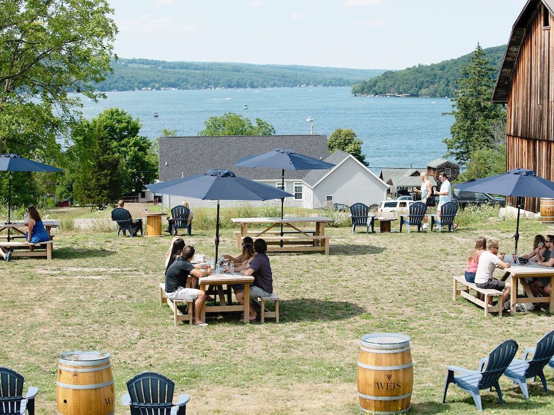 Guests relax at picnic tables and Adirondack chairs overlooking Keuka Lake at Weis Vineyards. Umbrellas provide shade while friends share wine and conversation near the rustic barn. The backdrop of water and hills makes this Finger Lakes winery a perfect summer destination.