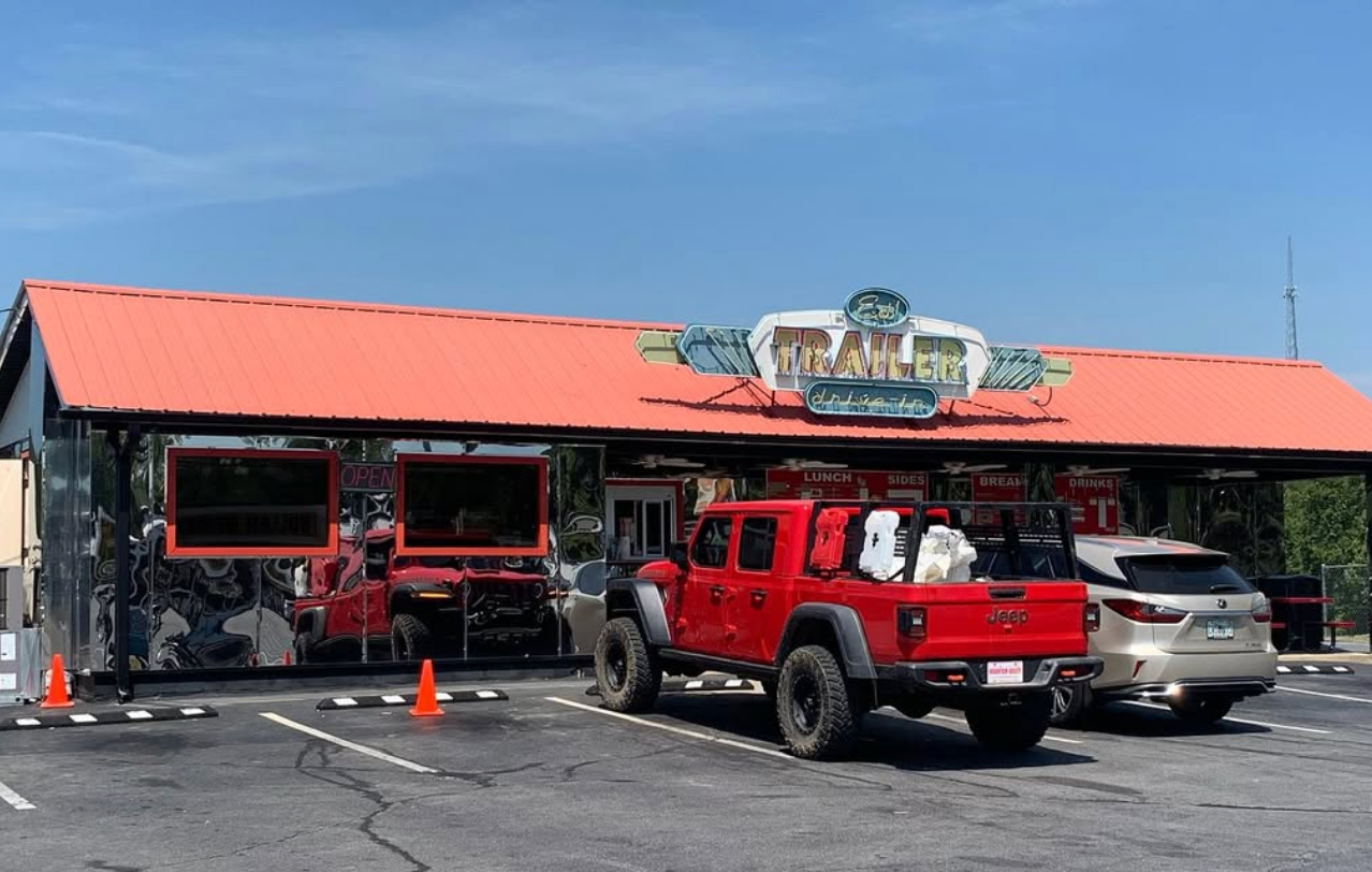 The exterior of The Trailer Drive-In featuring a bright red metal roof, reflective front windows, and a retro-style sign, with a red Jeep pickup and a silver SUV parked out front under a clear blue sky.