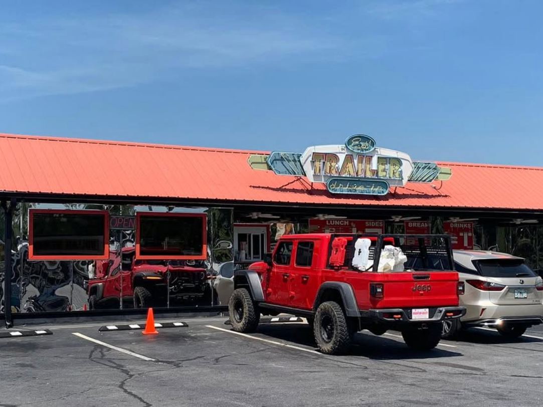 The exterior of The Trailer Drive-In featuring a bright red metal roof, reflective front windows, and a retro-style sign, with a red Jeep pickup and a silver SUV parked out front under a clear blue sky.