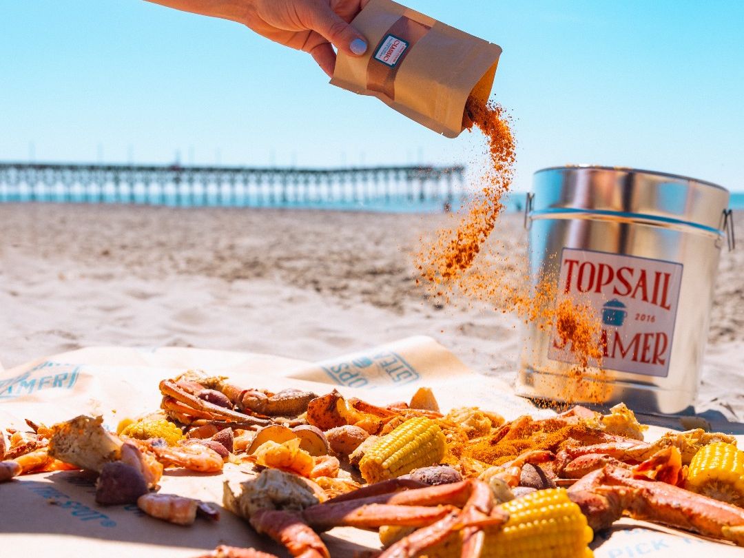 A hand sprinkles seasoning over a beachside seafood boil with crab legs and corn. Topsail Steamer brings delicious flavors to the sand with a casual, toes-in-the-sand dining vibe.