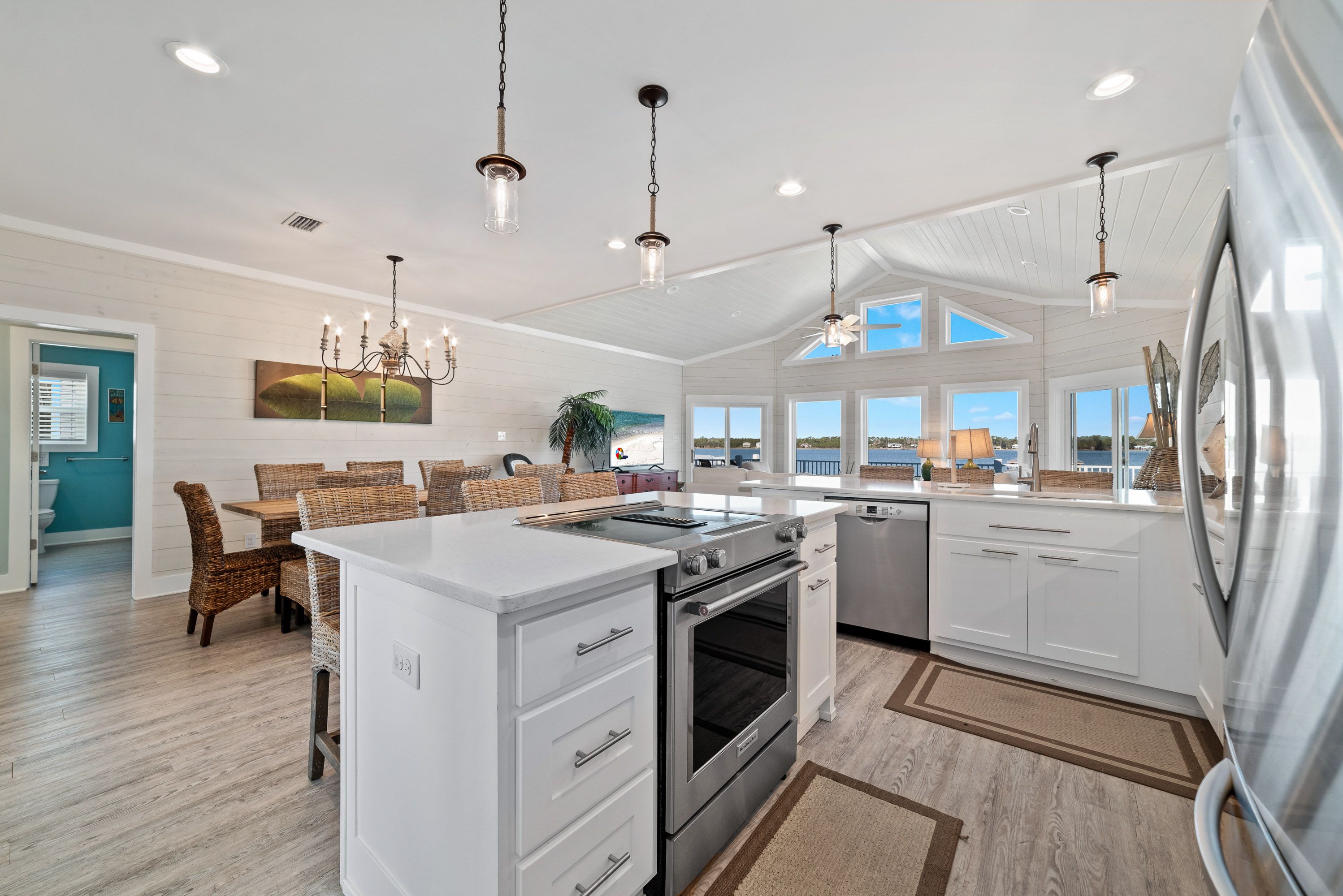 A bright coastal kitchen and dining area featuring white cabinetry, stainless steel appliances, wicker dining chairs, and vaulted ceilings with large windows offering panoramic water views.