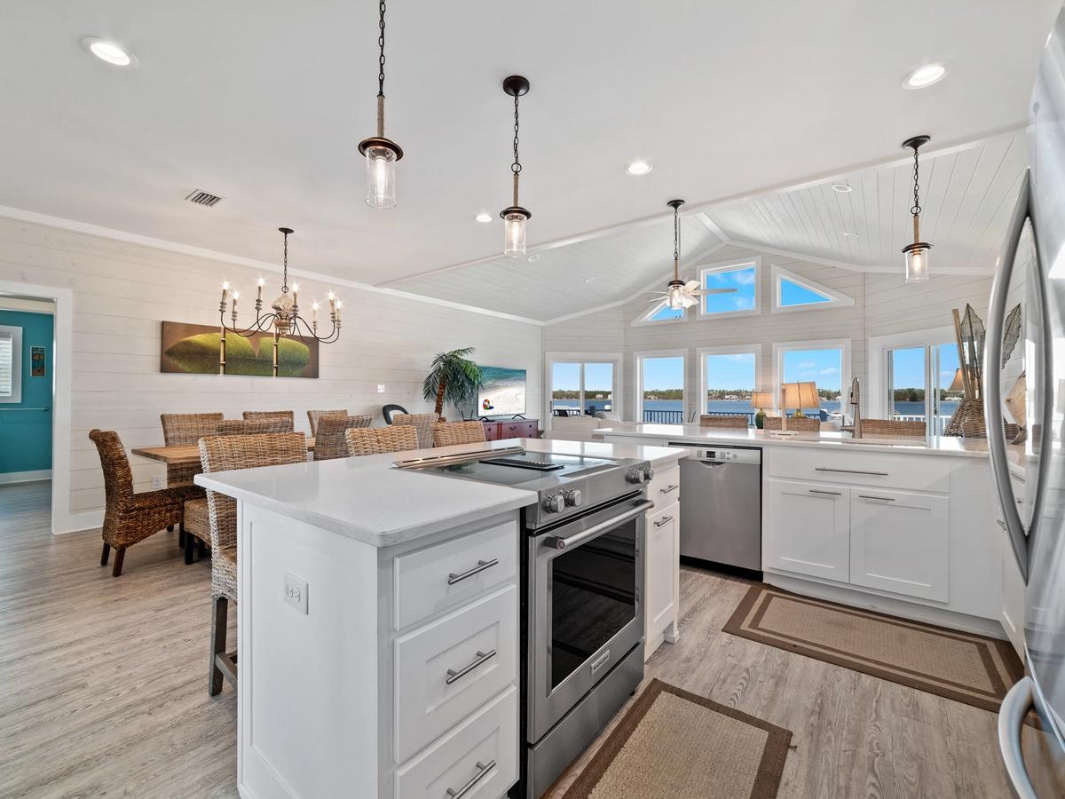 A bright coastal kitchen and dining area featuring white cabinetry, stainless steel appliances, wicker dining chairs, and vaulted ceilings with large windows offering panoramic water views.