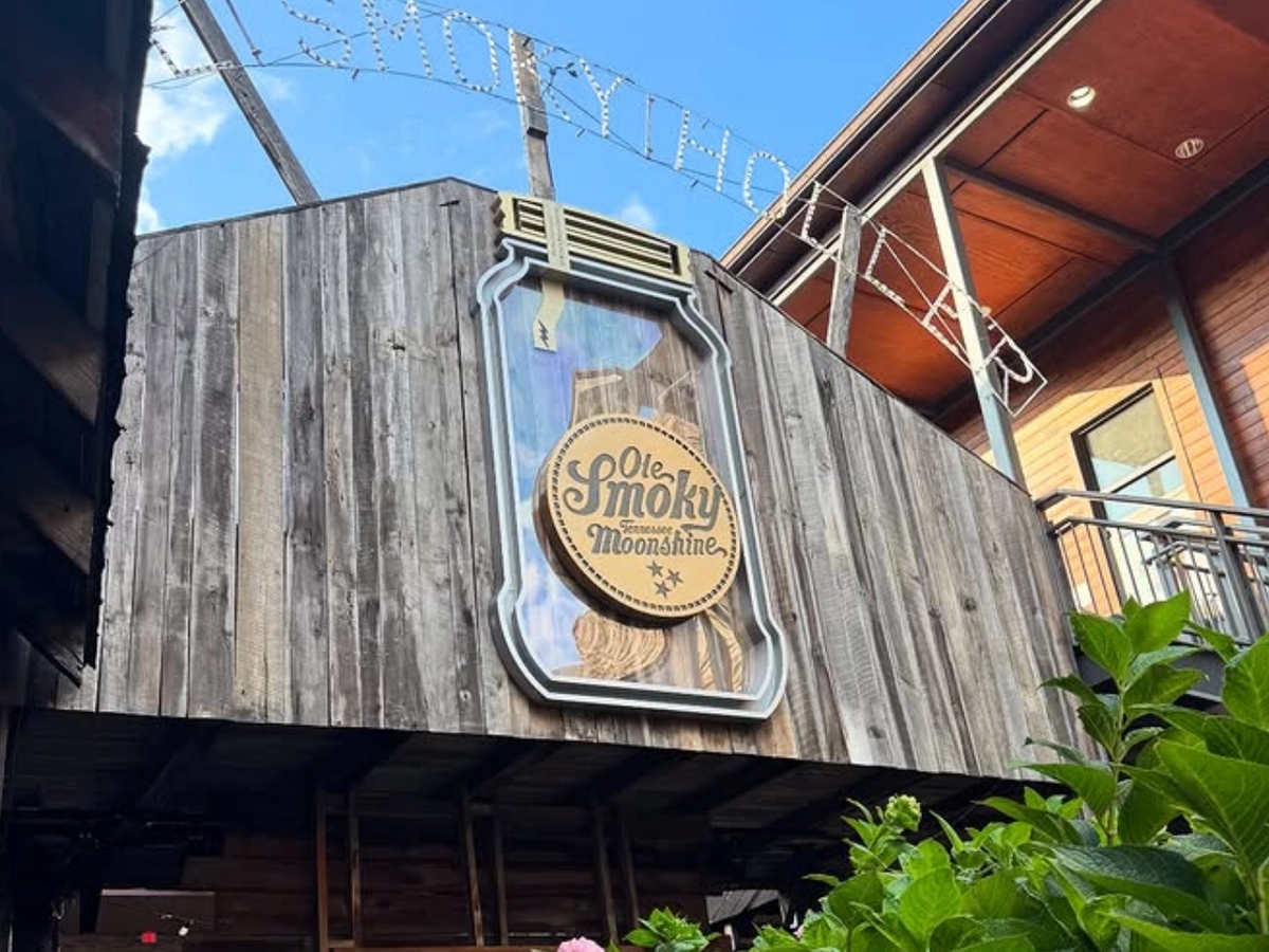 Wooden exterior of Ole Smoky Tennessee Moonshine distillery, featuring a large mason jar sign with the logo, framed by rustic architecture and greenery in the foreground.