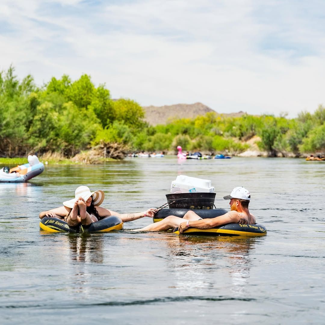 A couple floats lazily down a calm river in inflatable tubes, holding hands under the warm desert sun. Surrounded by greenery and mountains, it’s the perfect summer escape.