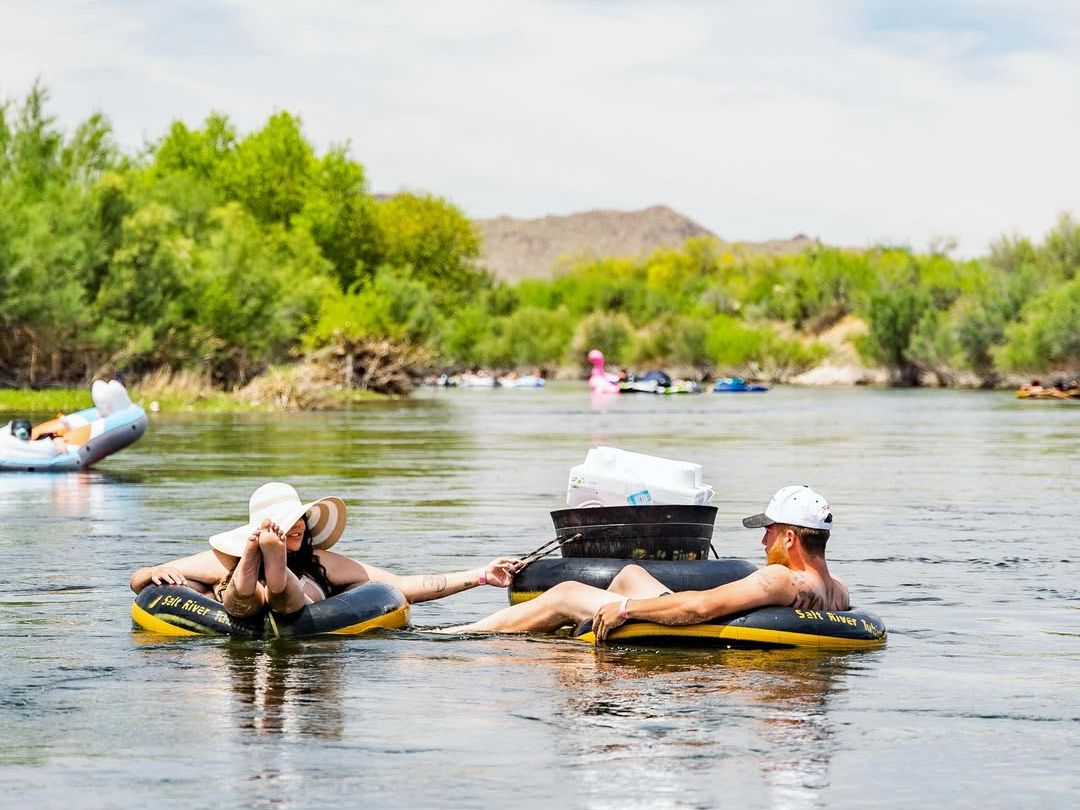 A couple floats lazily down a calm river in inflatable tubes, holding hands under the warm desert sun. Surrounded by greenery and mountains, it’s the perfect summer escape.