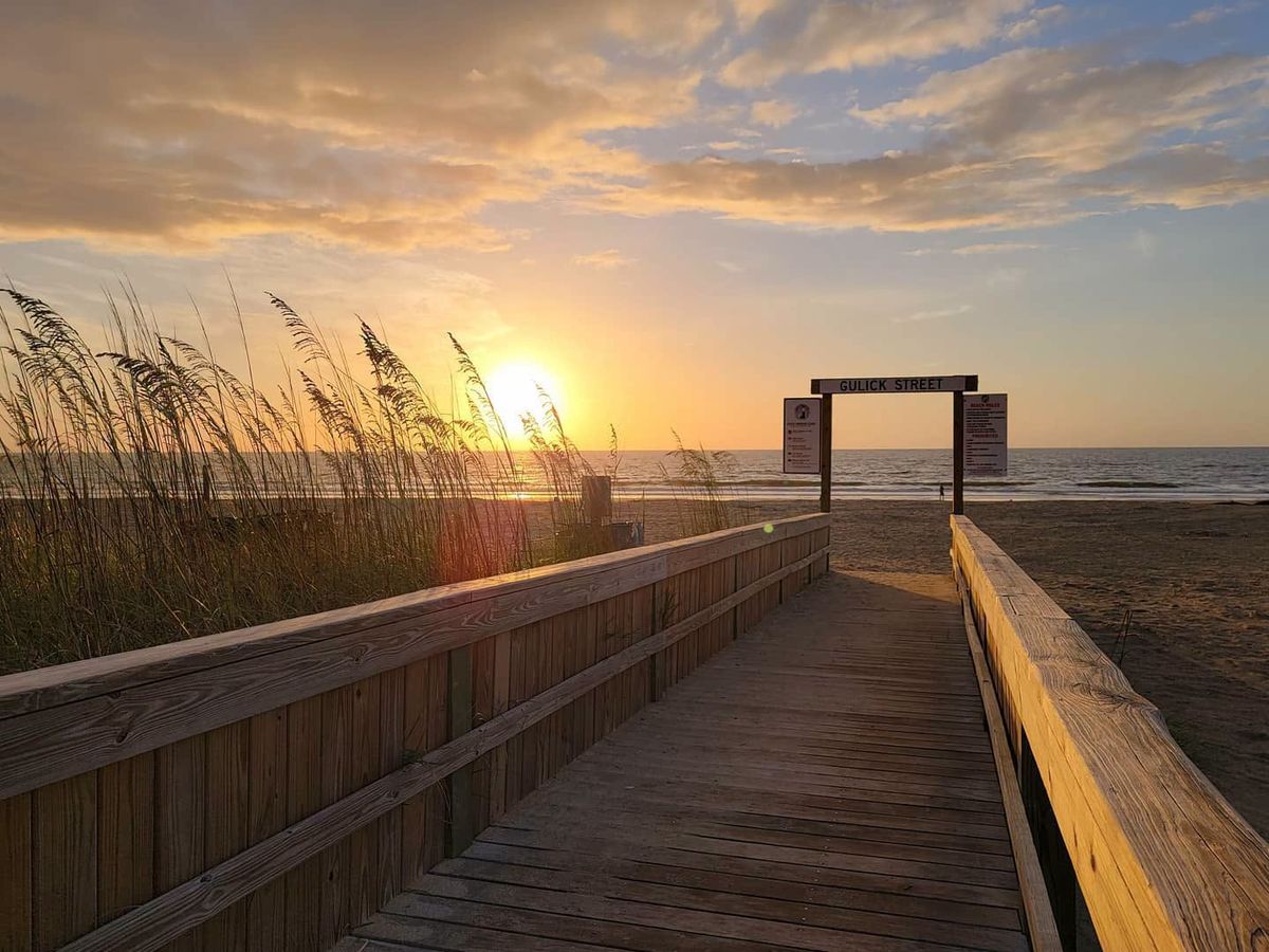 A weathered wooden boardwalk leads toward the beach beneath a radiant sunrise, framed by golden sea oats swaying in the breeze. A "Gulick Street" beach access sign marks the entrance as the sun rises over the ocean. 