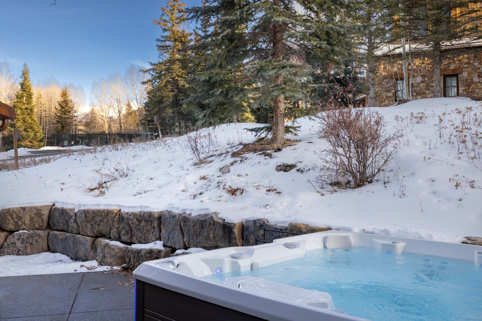 Outdoor hot tub steaming in a snowy mountain setting, nestled beside stone retaining walls and evergreen trees, offering a cozy winter soak with alpine lodge surroundings under clear blue skies.