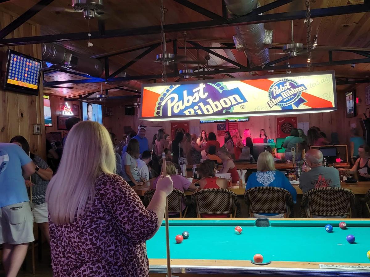A woman holds a pool cue in the foreground of a lively, wood-paneled bar and grill, where a crowd gathers beneath a glowing Pabst Blue Ribbon light fixture above the billiard table. Patrons fill the bar seating and dartboards line the back wall in the warm, energetic space. 