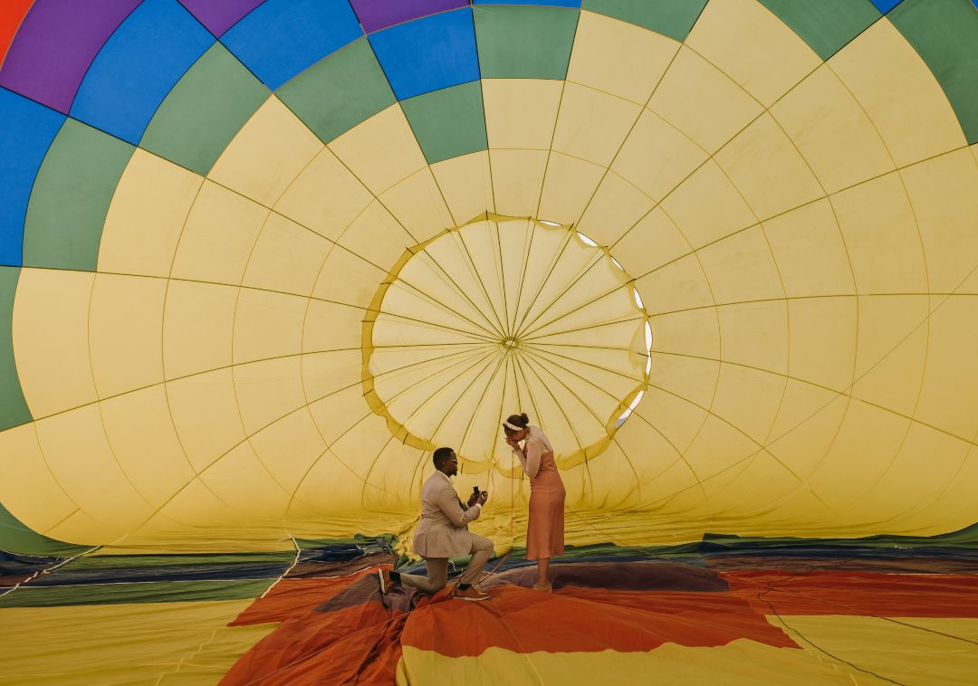 A man kneels to propose inside a colorful hot air balloon before takeoff, surrounded by glowing yellow and rainbow panels. The romantic moment captures love and adventure in perfect harmony.