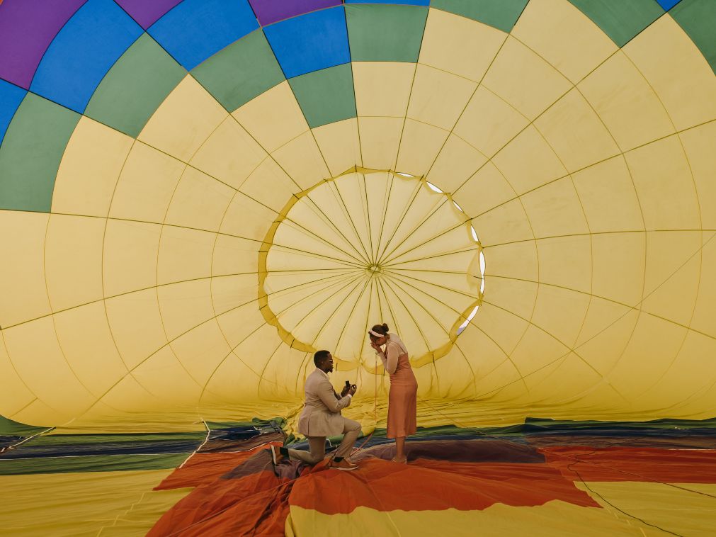 A man kneels to propose inside a colorful hot air balloon before takeoff, surrounded by glowing yellow and rainbow panels. The romantic moment captures love and adventure in perfect harmony.