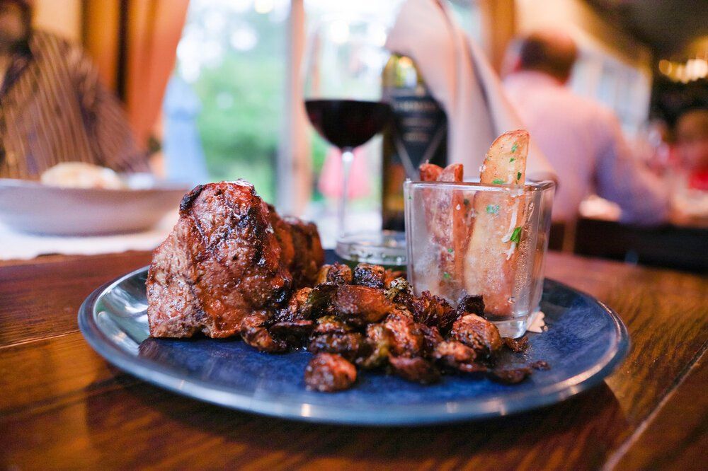 A beautifully plated brunch spread with eggs Benedict, croissants, coffee, and fresh drinks sits on a wooden table. Large windows let in natural light, creating a warm and welcoming dining space. Lakeside Restaurant is known for its elegant brunch and scenic setting.