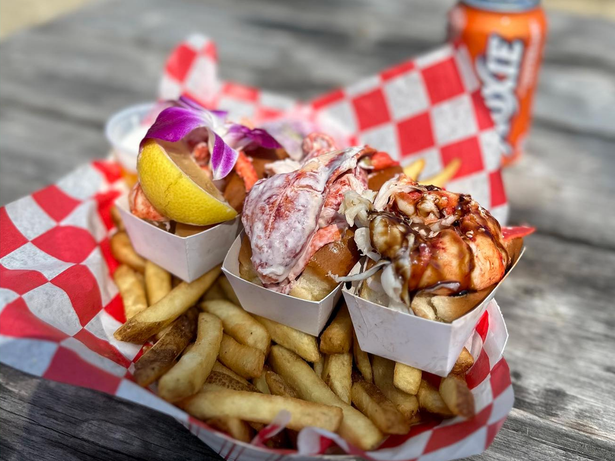 A trio of gourmet lobster rolls topped with lemon, sauce, and edible flowers is served over a bed of golden fries in a red checkered basket, with a can of orange soda in the background.