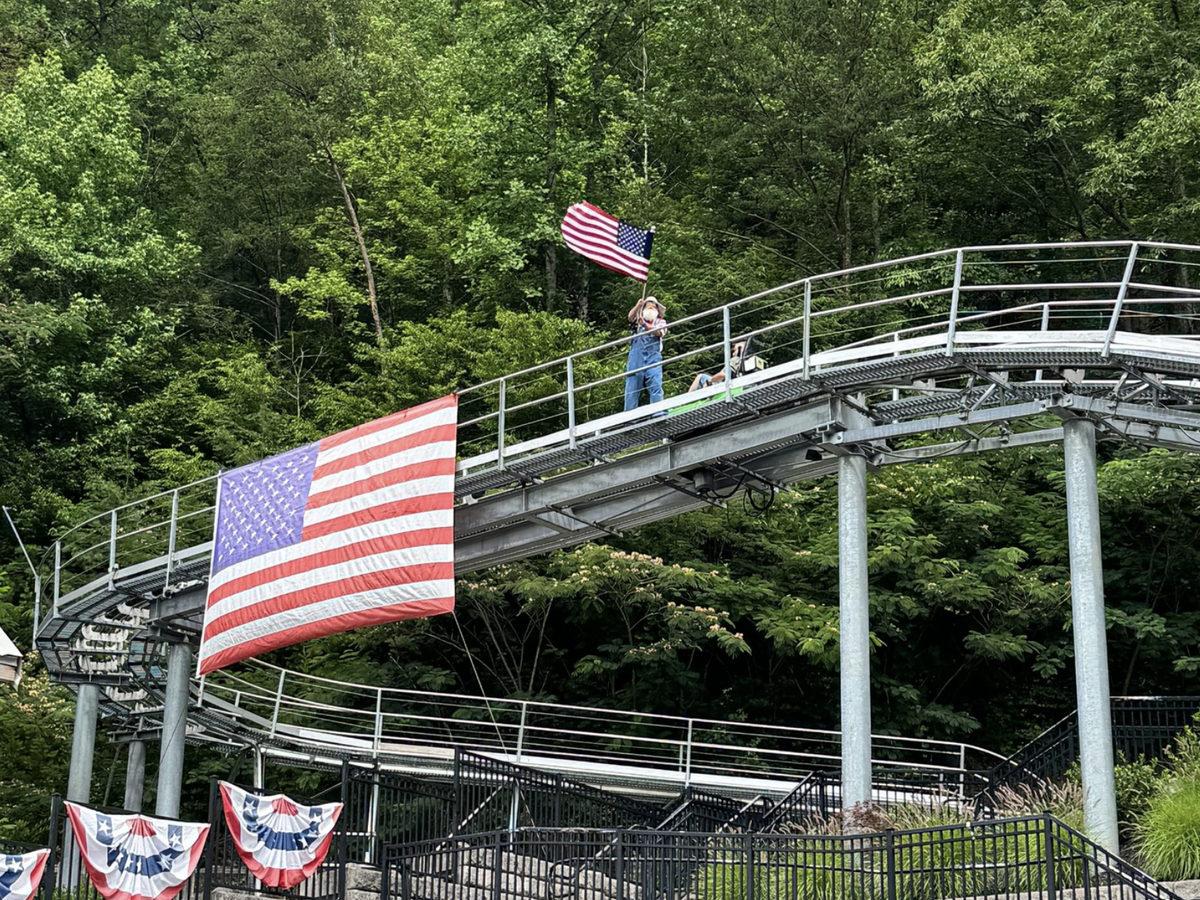 A person in overalls waves an American flag while standing on a mountain coaster track, decorated with large U.S. flags and patriotic bunting, surrounded by dense green forest.