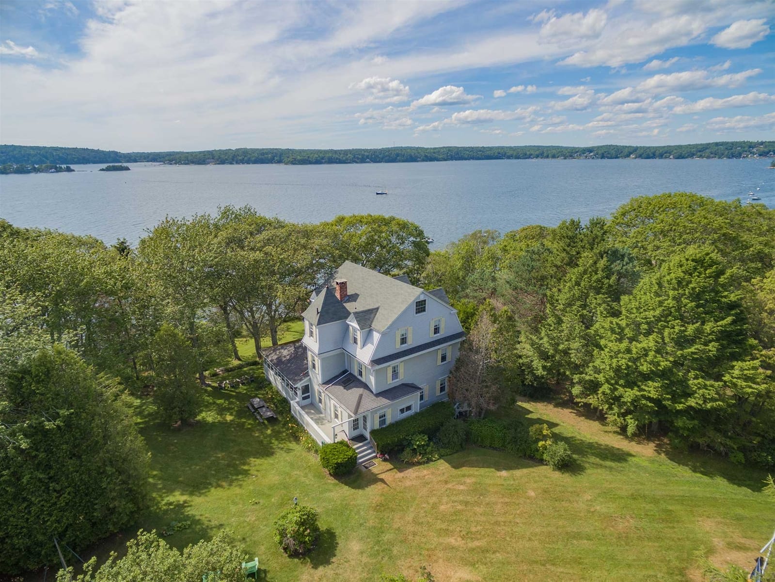 A large coastal home with gray siding and white trim sits on a grassy lot surrounded by trees, overlooking a calm blue lake under a bright sky with scattered clouds.