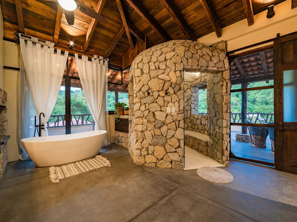 This rustic-chic bathroom features stone walls, wood ceilings, and a deep soaking tub with jungle views. It’s a spa-like retreat tucked in a luxurious tropical escape.