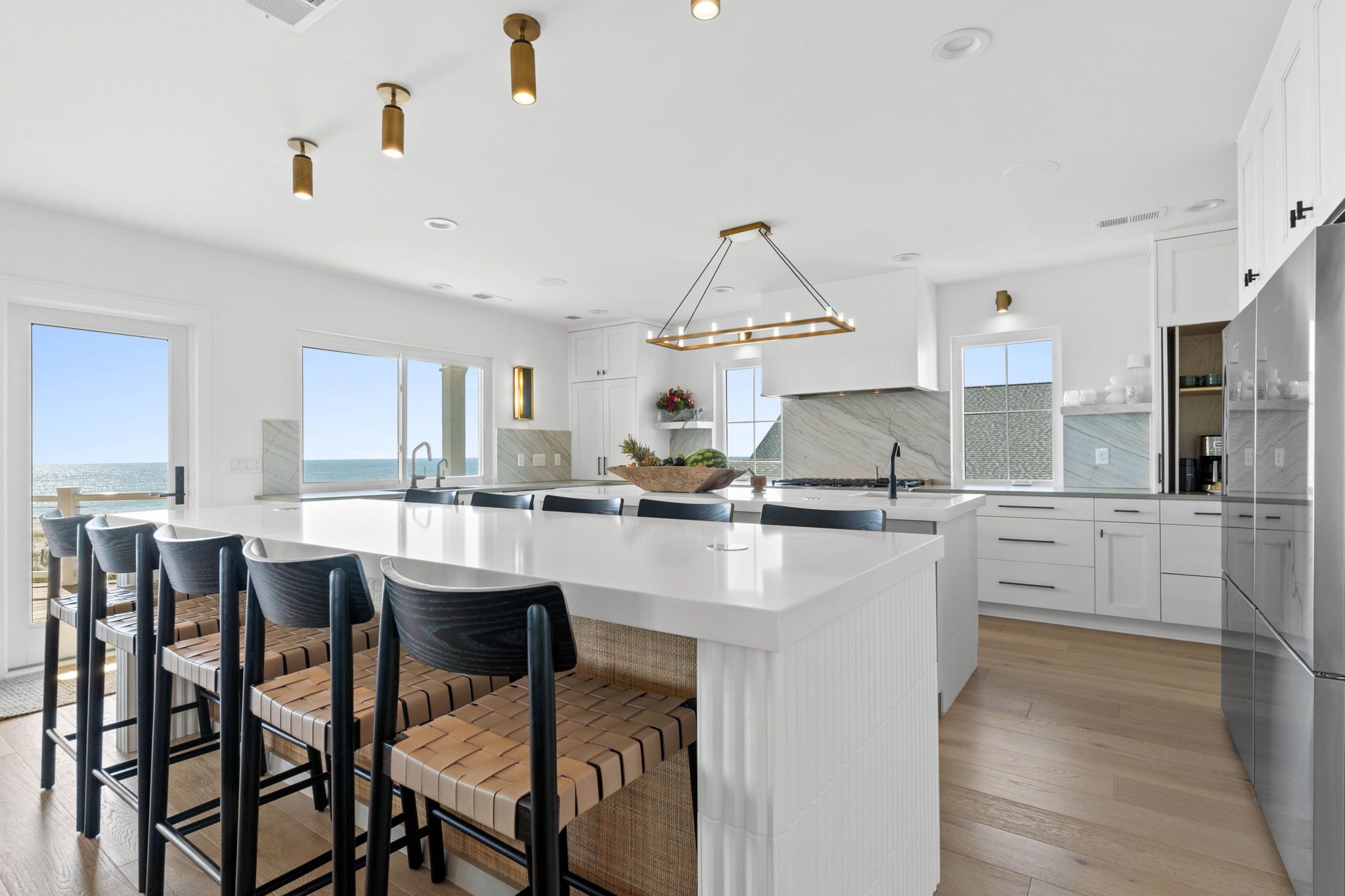 Bright coastal kitchen with a large white island, ocean views, woven barstools, and gold-accented lighting