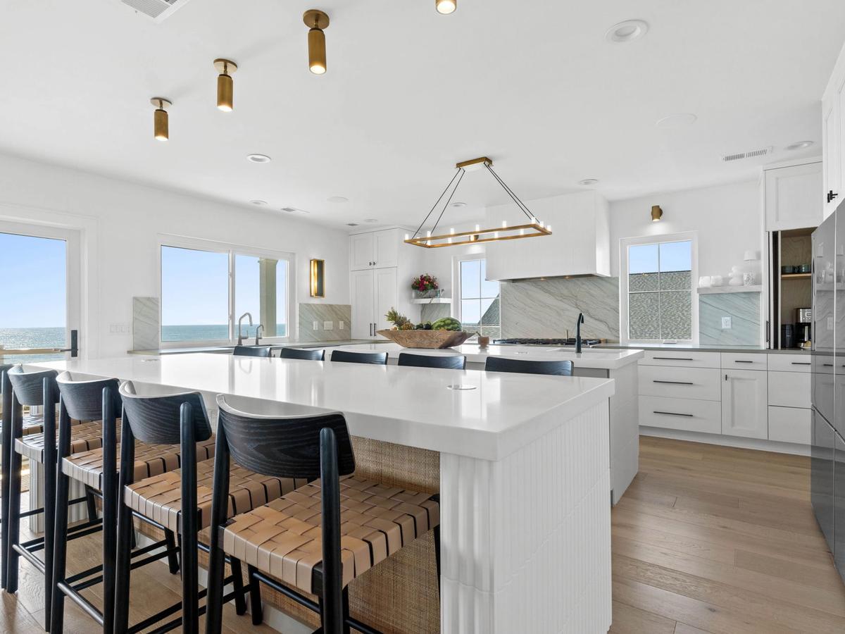 Bright coastal kitchen with a large white island, ocean views, woven barstools, and gold-accented lighting