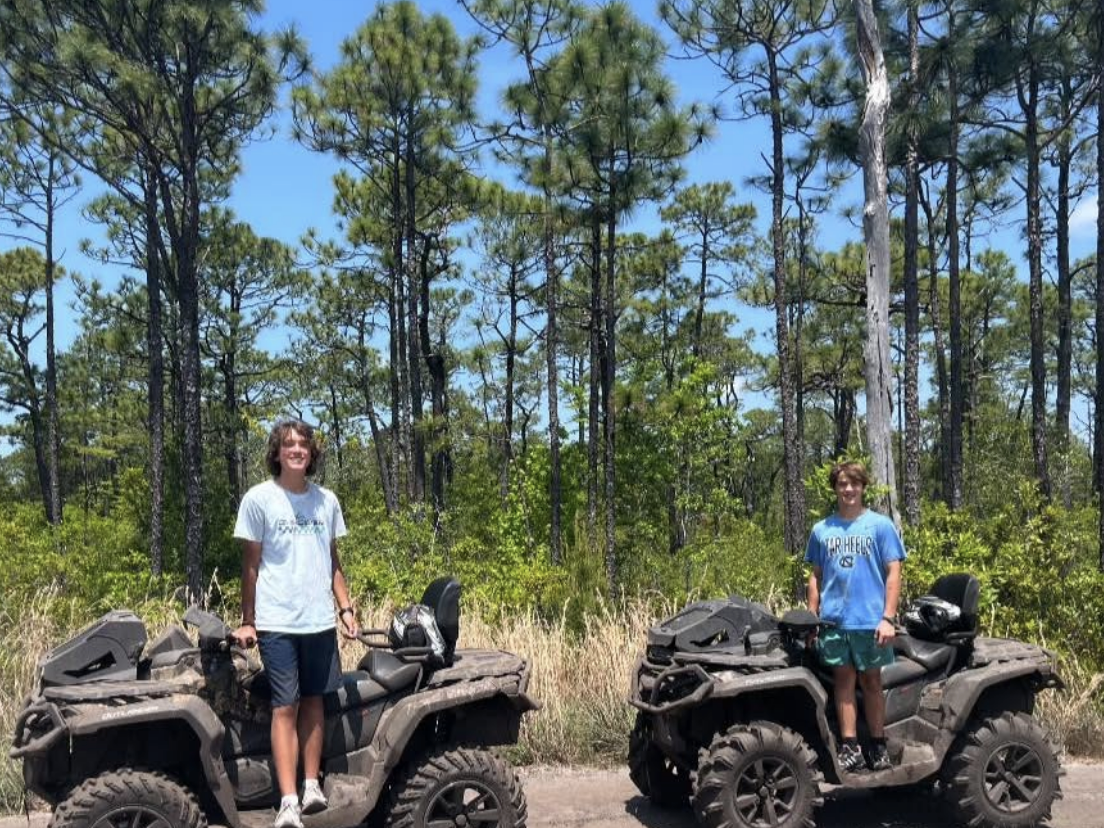 Two people standing on muddy ATVs in front of a pine forest on a sunny day, smiling and enjoying an off-road adventure.