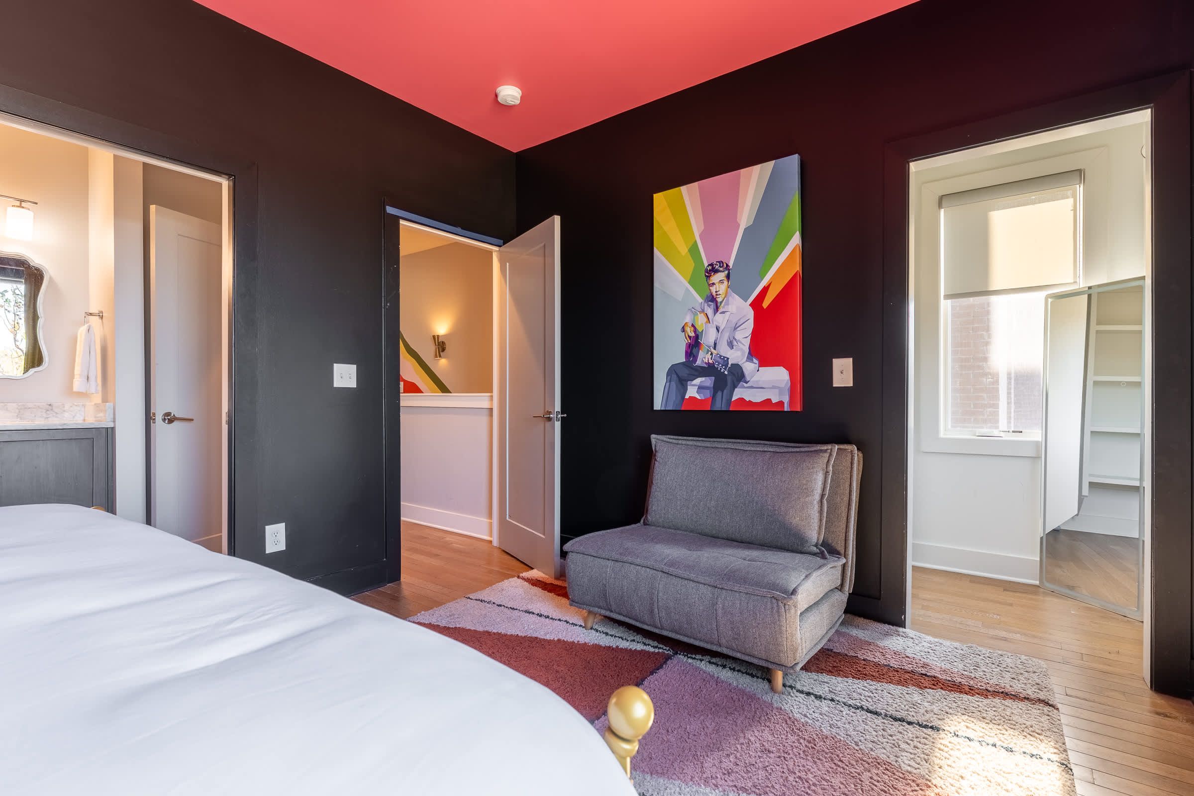 A bold, music-inspired bedroom with black walls, a red ceiling, hardwood floors, a cozy gray lounge chair, and vibrant Elvis artwork creating a dramatic yet inviting Nashville retreat.