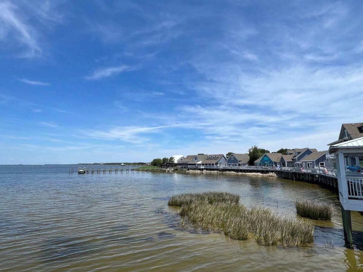 wooden boardwalk lined with blue and gray coastal cottages and shops curves along the calm sound waters of the Southern Outer Banks. Marsh grasses grow in the shallows beneath a wide sky streaked with wispy clouds