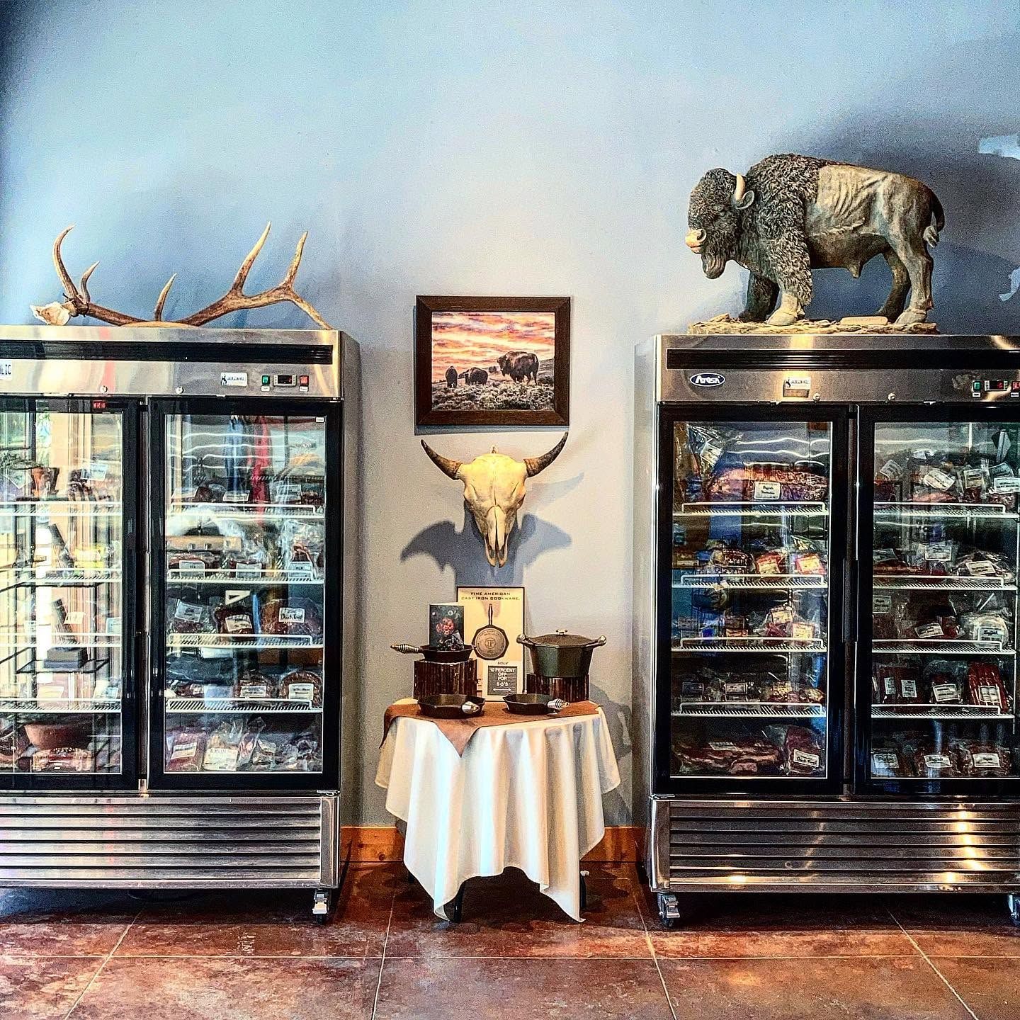 Two glass-front coolers packed with vacuum-sealed jerky and specialty meats flank a small display table of cast iron cookware at Jackson Hole Buffalo Meat Co. A mounted bison sculpture, elk antlers, a bison skull, and a framed prairie painting reinforce the shop's Western character. 
