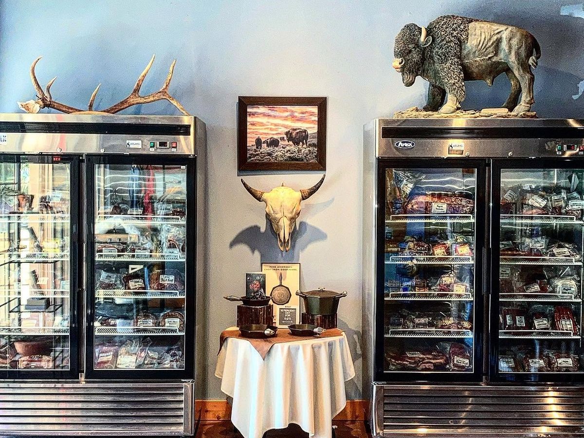 Two glass-front coolers packed with vacuum-sealed jerky and specialty meats flank a small display table of cast iron cookware at Jackson Hole Buffalo Meat Co. A mounted bison sculpture, elk antlers, a bison skull, and a framed prairie painting reinforce the shop's Western character.