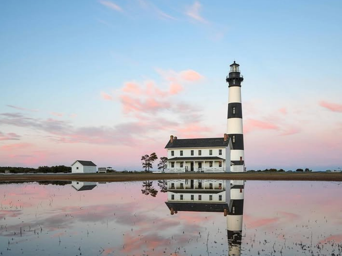 A black-and-white lighthouse and keeper’s house reflect in still water beneath a pastel pink and blue sunset sky.