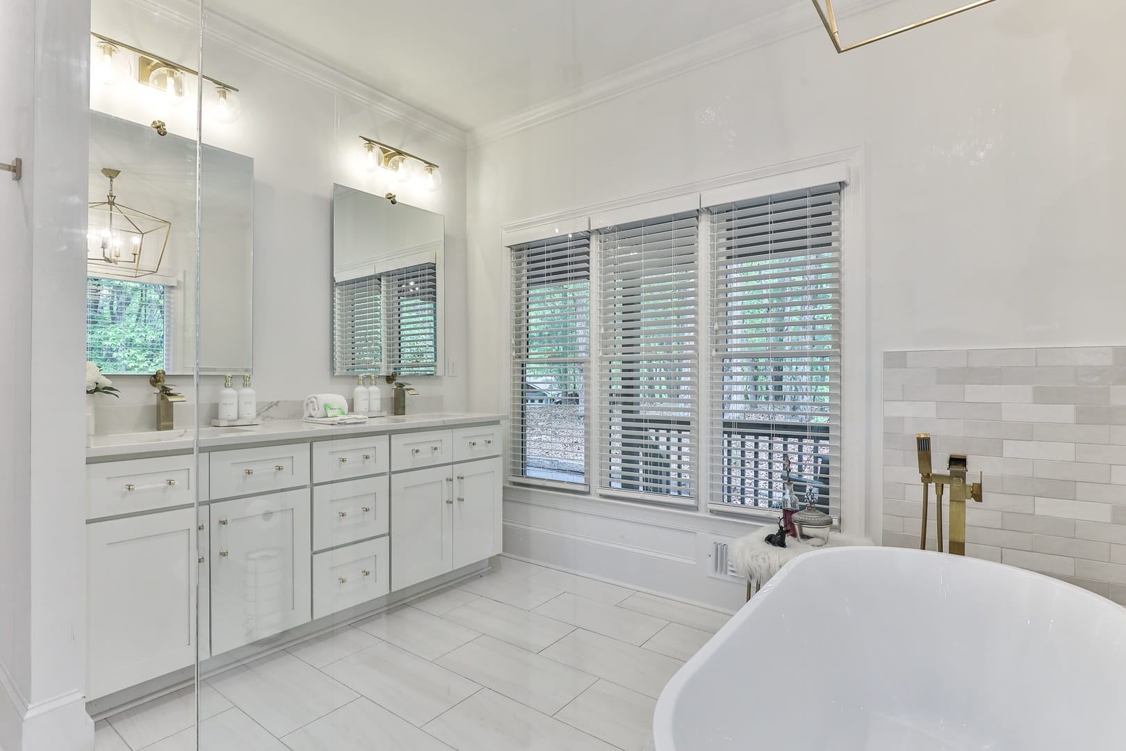 Elegant bathroom with white cabinetry, double sinks with mirrors, a freestanding bathtub, and large windows with blinds letting in natural light.