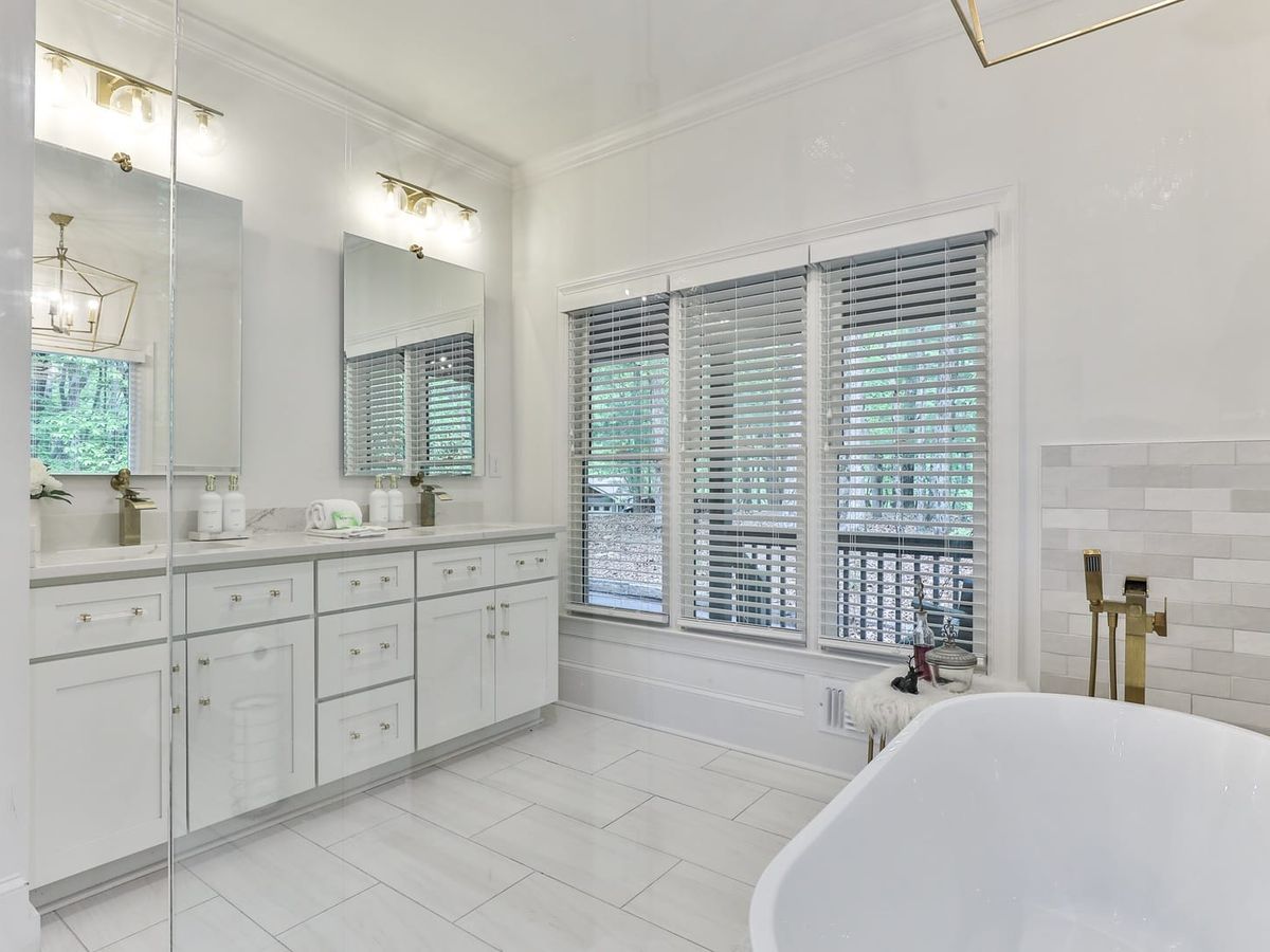 Elegant bathroom with white cabinetry, double sinks with mirrors, a freestanding bathtub, and large windows with blinds letting in natural light.