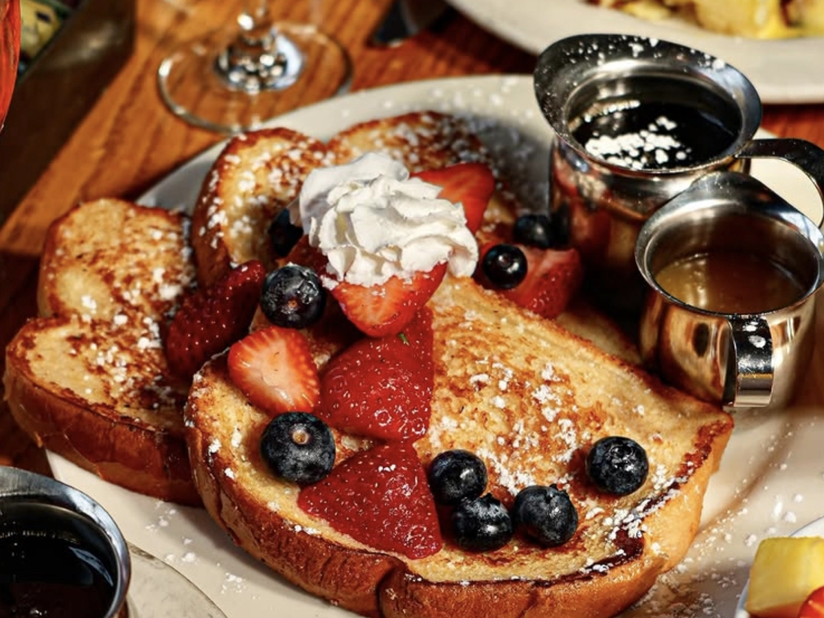 Thick-cut French toast topped with fresh strawberries, blueberries, a dollop of whipped cream, and a dusting of powdered sugar, served with maple syrup and a side of warm caramel sauce.