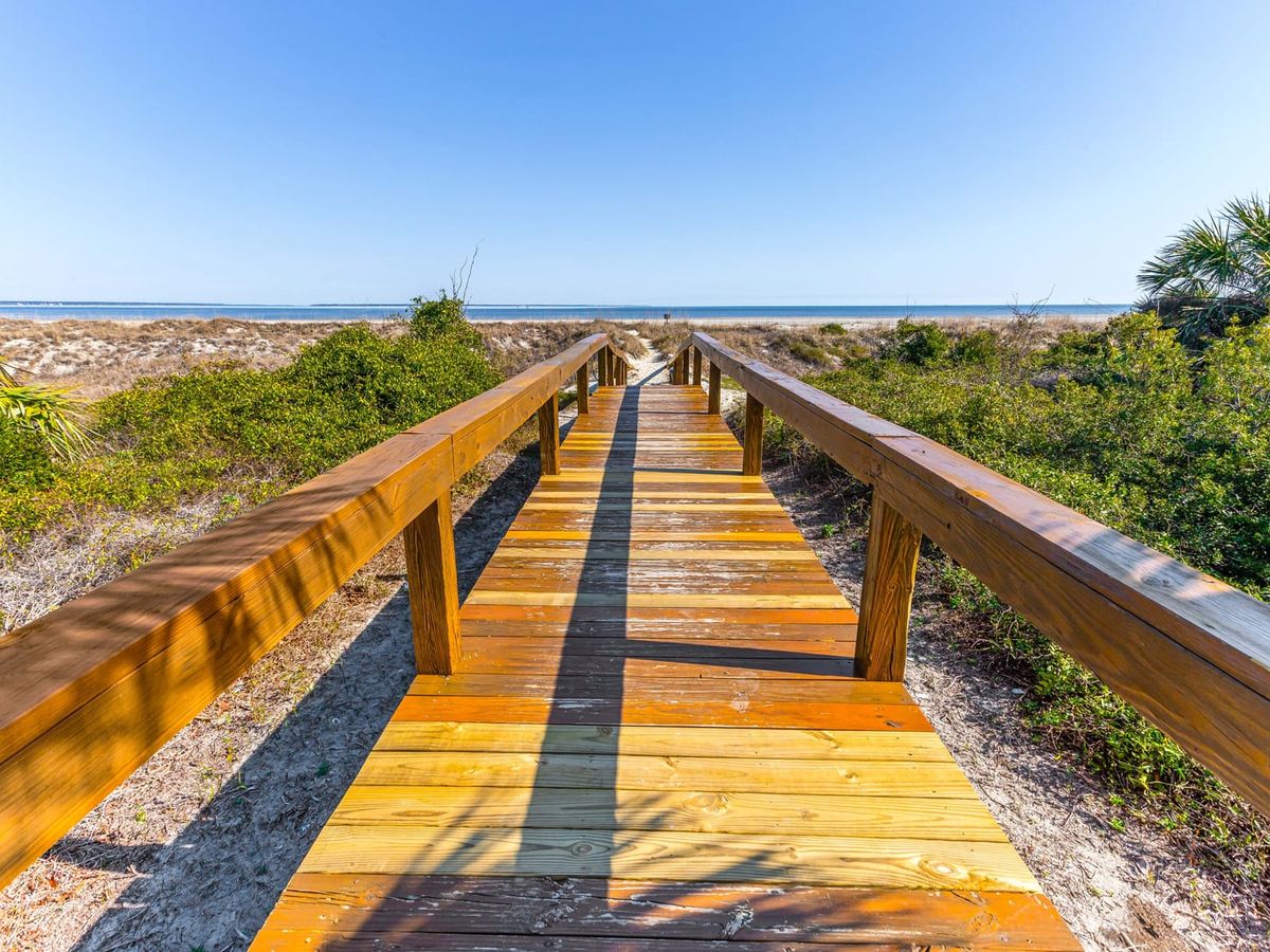 A weathered wooden boardwalk leads through coastal dunes and sea grasses toward the open ocean on Tybee Island, framed by palmettos and shrubs under a clear blue sky. The elevated walkway provides easy beach access while protecting the natural dune ecosystem.