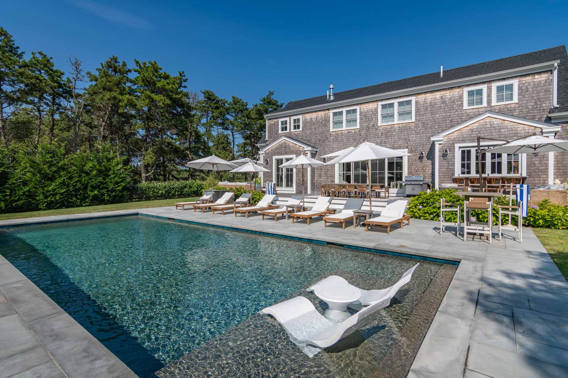 A scenic waterfront home sits on rocky shoreline surrounded by trees and ocean views in Martha’s Vineyard. A private dock stretches into the calm water, creating a peaceful coastal setting. This beautiful New England destination is known for charming homes and stunning seaside landscapes.