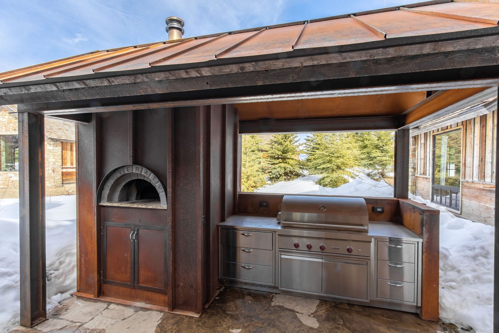A covered outdoor mountain kitchen with a built-in stainless steel grill and wood-fired pizza oven, framed by rustic metal and wood finishes and surrounded by a snowy alpine landscape.