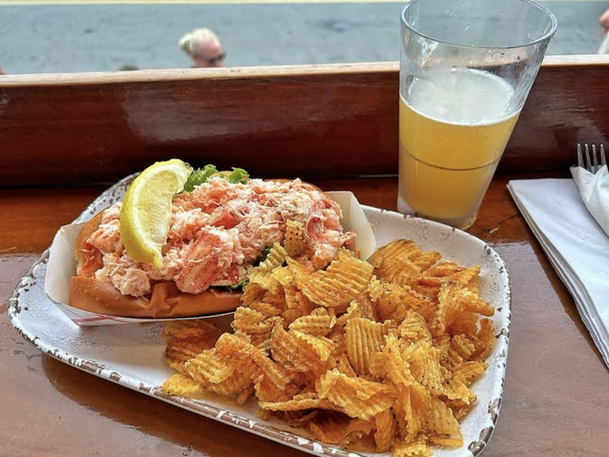 Tray with a lobster roll topped with a lemon wedge, a pile of crinkle-cut potato chips, and a glass of beer, set on a wooden counter by a window