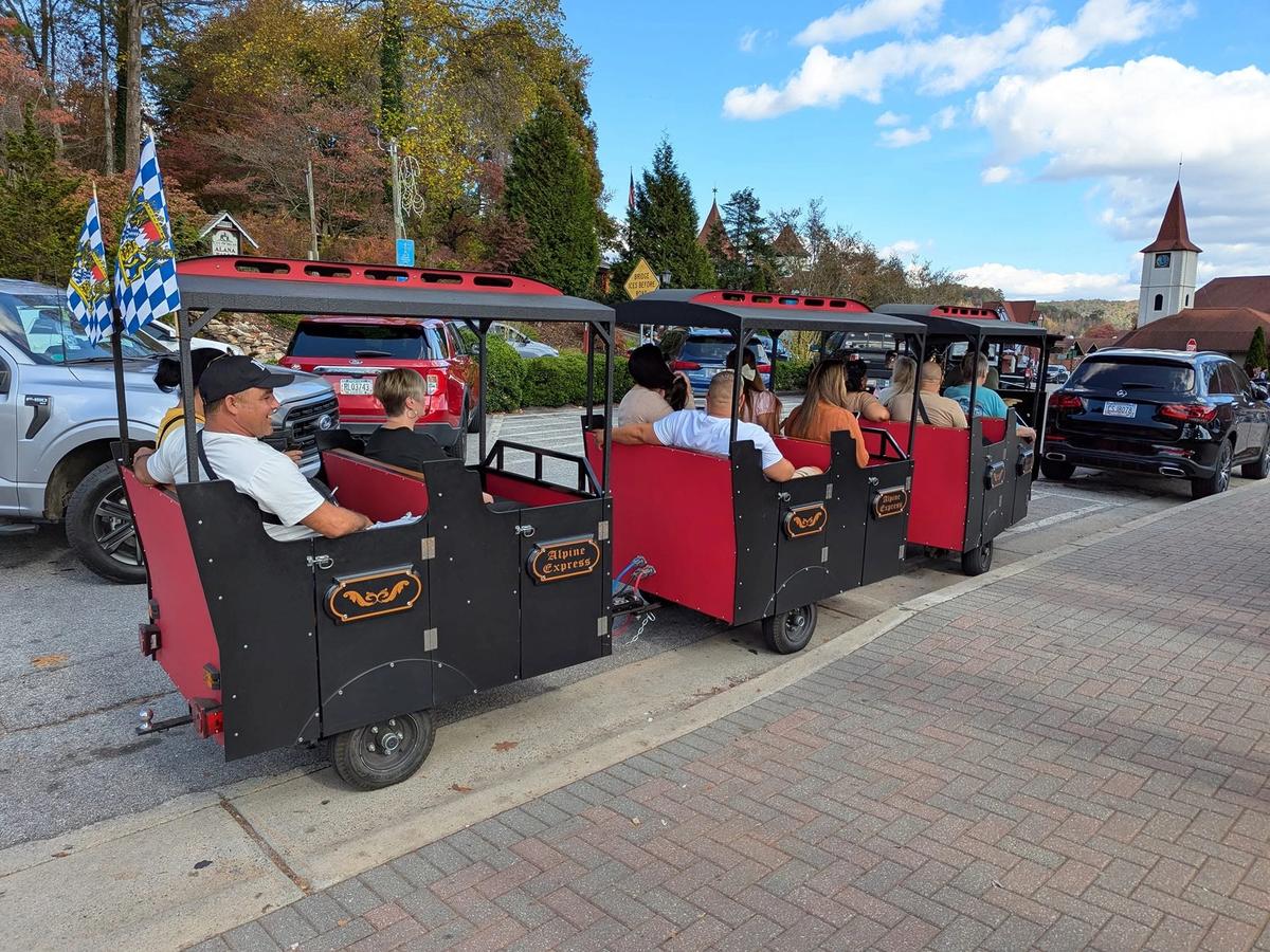 The Alpine Express tourist trolley carries visitors through the charming Bavarian-style streets of Helen, Georgia, during the Christkindlmarkt Alpine holiday market, with a steepled church and fall foliage visible in the background.