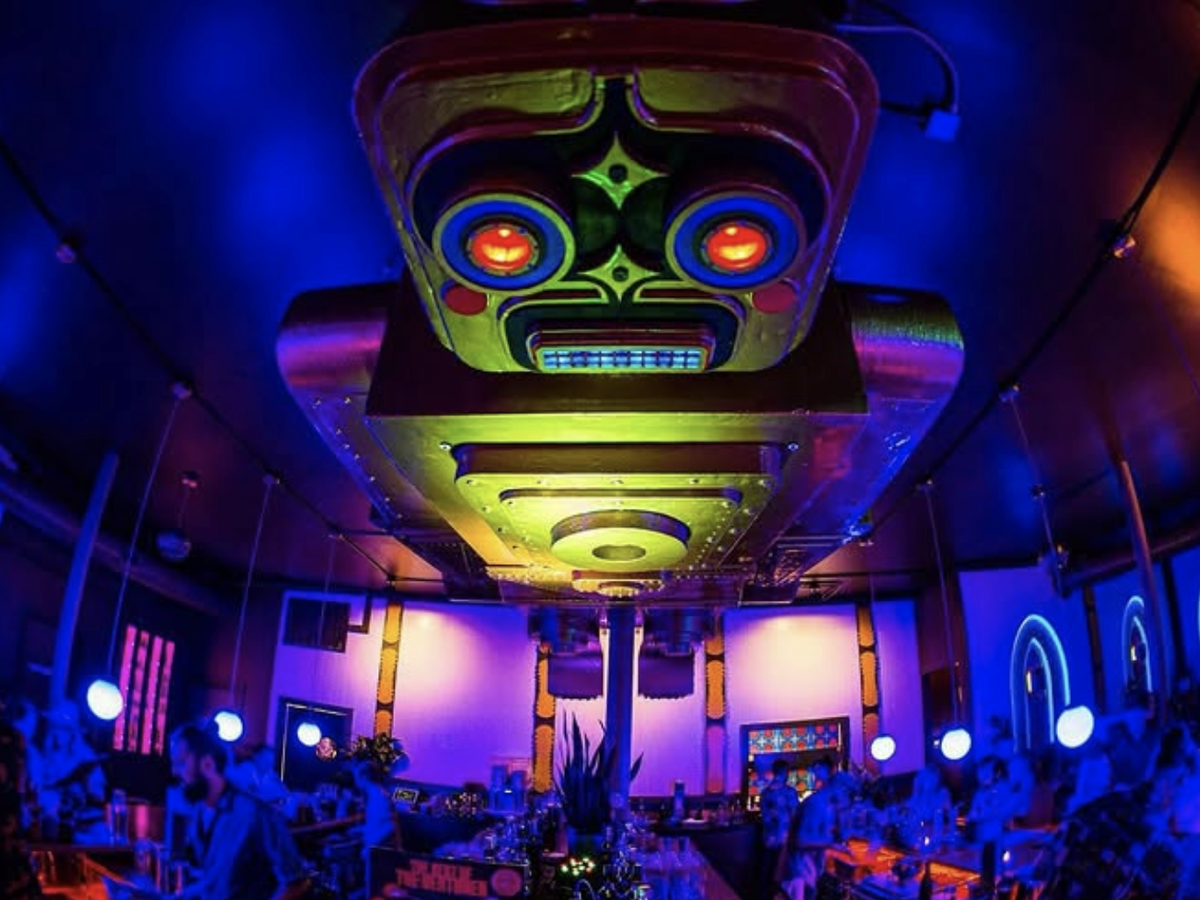 A futuristic bar with neon lighting and a giant glowing robot face installation on the ceiling above the crowd.