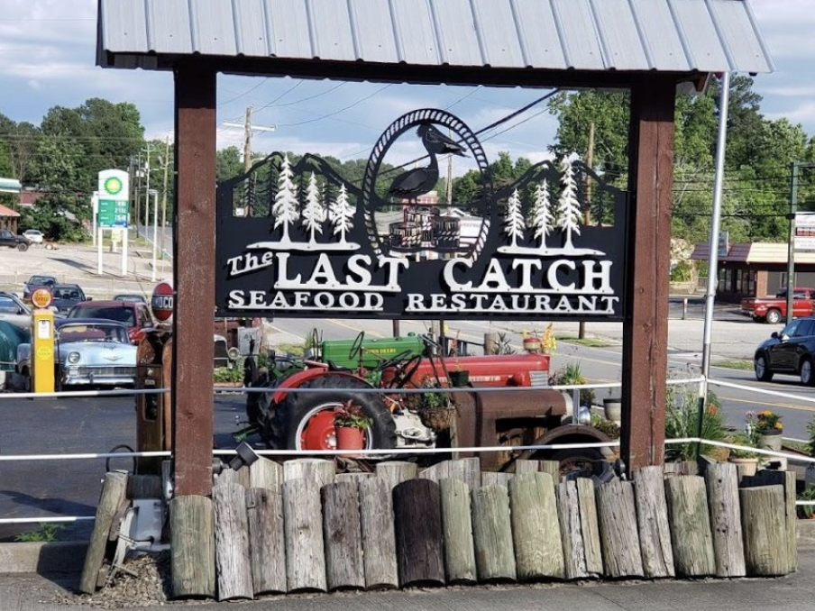 A rustic roadside sign for The Last Catch Seafood Restaurant stands under a metal roof with a carved wooden design featuring a fish, trees, and mountains, set against a backdrop of vintage cars and a BP gas station.