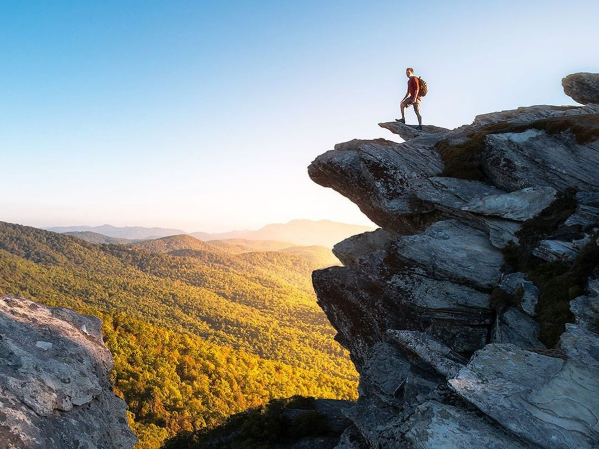 A hiker stands on a dramatic rocky outcrop at Grandfather Mountain, overlooking rolling forested hills glowing in golden sunlight under a clear blue sky.