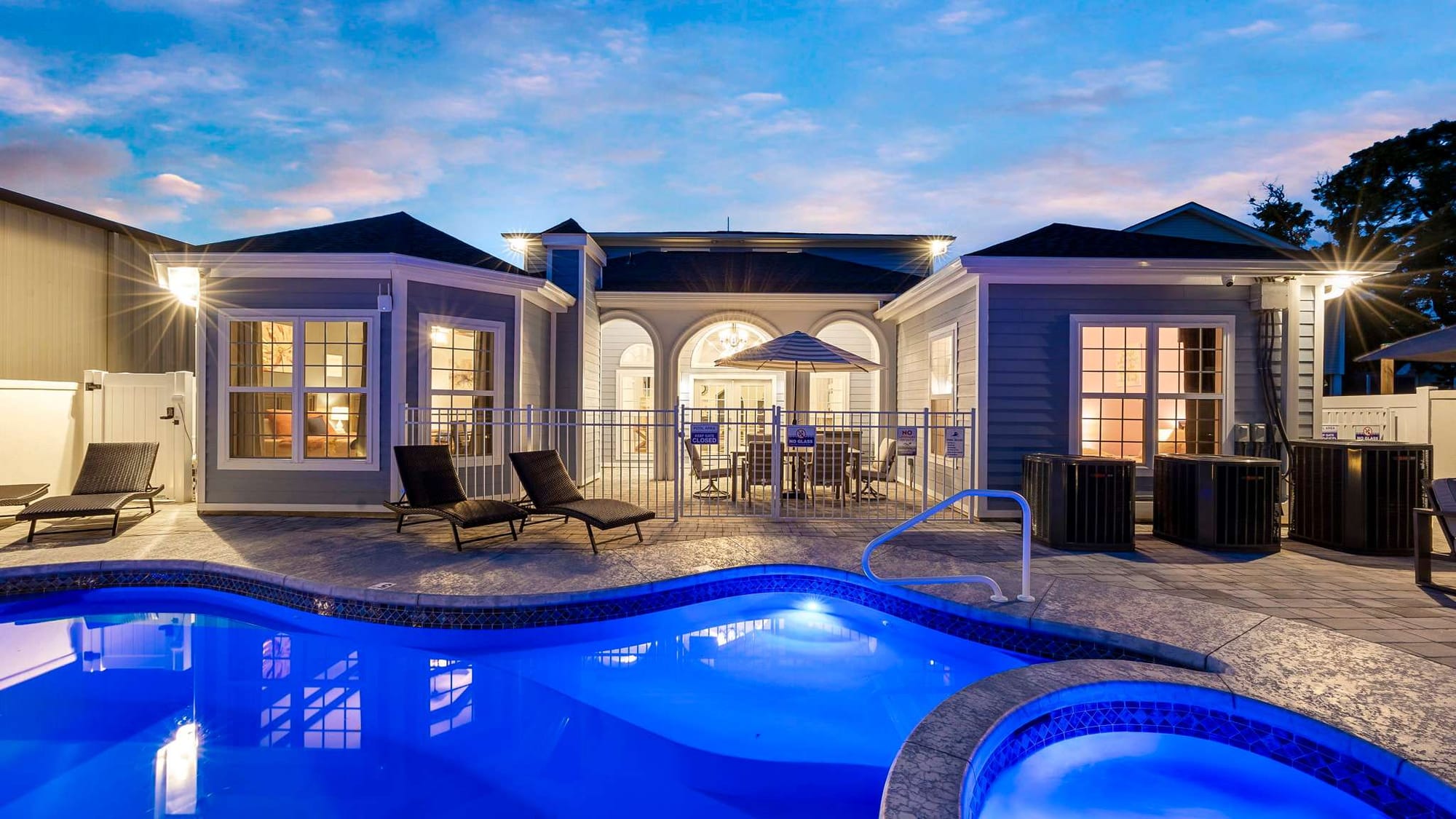 A luxury vacation home clubhouse at dusk with a glowing resort-style swimming pool and hot tub in the foreground, lounge chairs along the patio, and warmly lit interiors framed by arched windows under a soft blue evening sky.