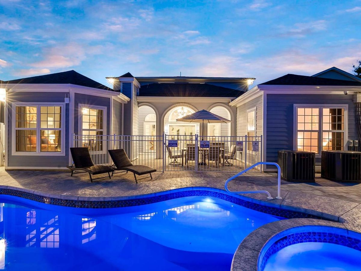 A luxury vacation home clubhouse at dusk with a glowing resort-style swimming pool and hot tub in the foreground, lounge chairs along the patio, and warmly lit interiors framed by arched windows under a soft blue evening sky.