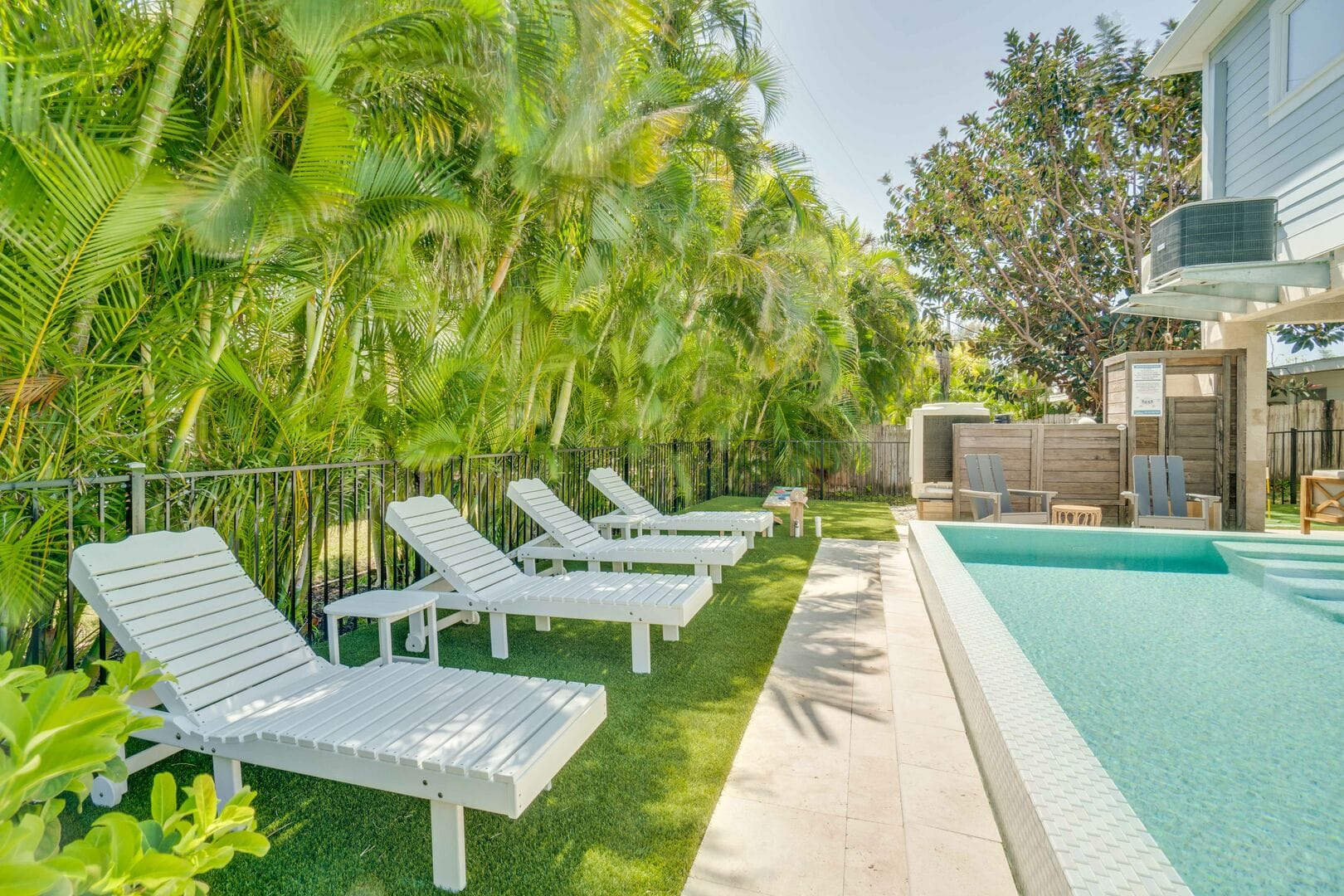 White lounge chairs line a sunlit poolside lawn bordered by lush tropical palms, creating a private, resort-style setting beside a sparkling turquoise pool.