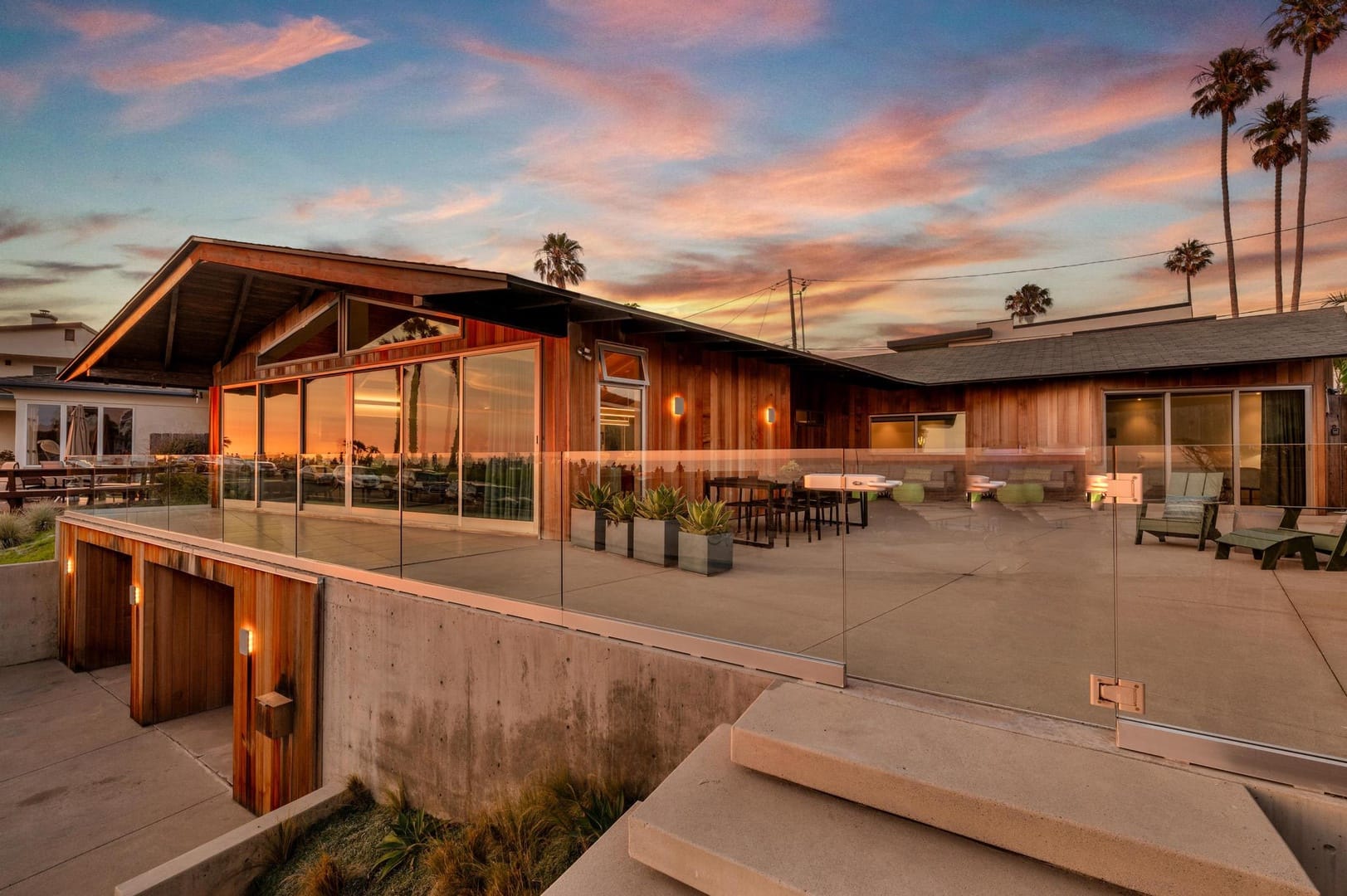 A modern coastal home in San Diego with glass walls and sleek wood details, overlooking a breathtaking sunset. The wide terrace and ocean backdrop make this a dream setting for evening gatherings.